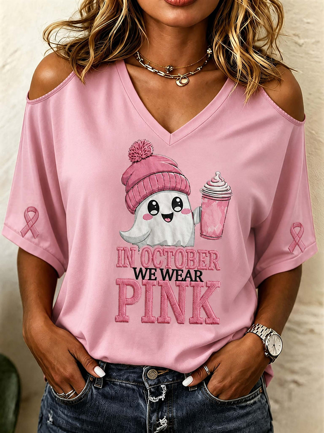 A woman wearing a pink cold-shoulder V-neck t-shirt with a cartoon ghost graphic holding a pink drink and the text 'IN OCTOBER WE WEAR PINK'.