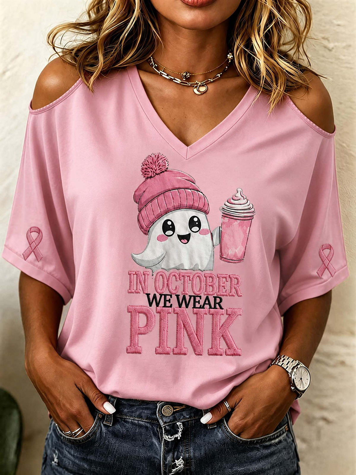 A woman wearing a pink cold-shoulder V-neck t-shirt with a cartoon ghost graphic holding a pink drink and the text 'IN OCTOBER WE WEAR PINK'.