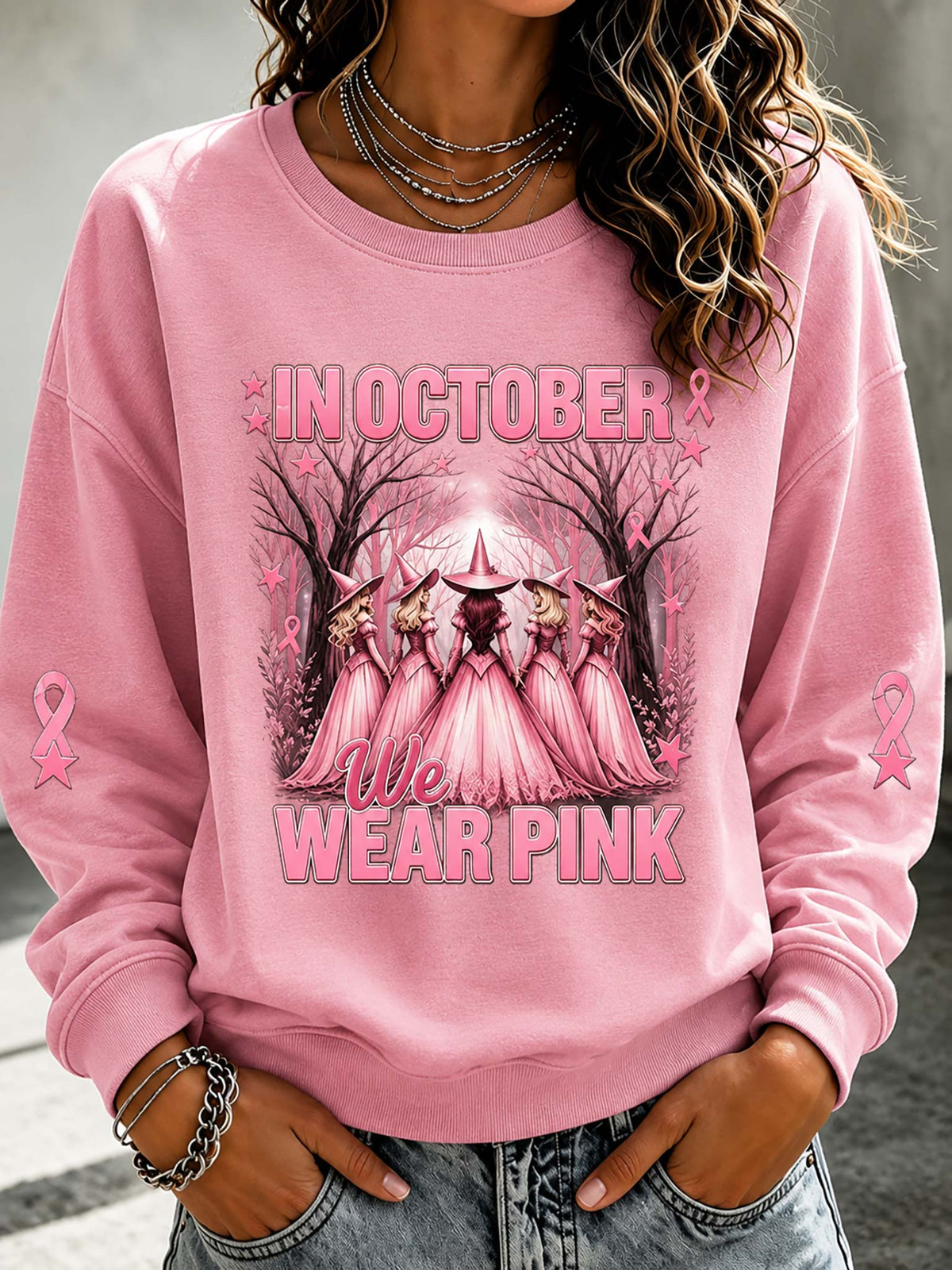 A woman wearing a pink crewneck sweatshirt with a graphic that reads 'IN OCTOBER WE WEAR PINK' and shows a group of women in pink dresses under a crescent moon, with pink awareness ribbons on the sleeves.