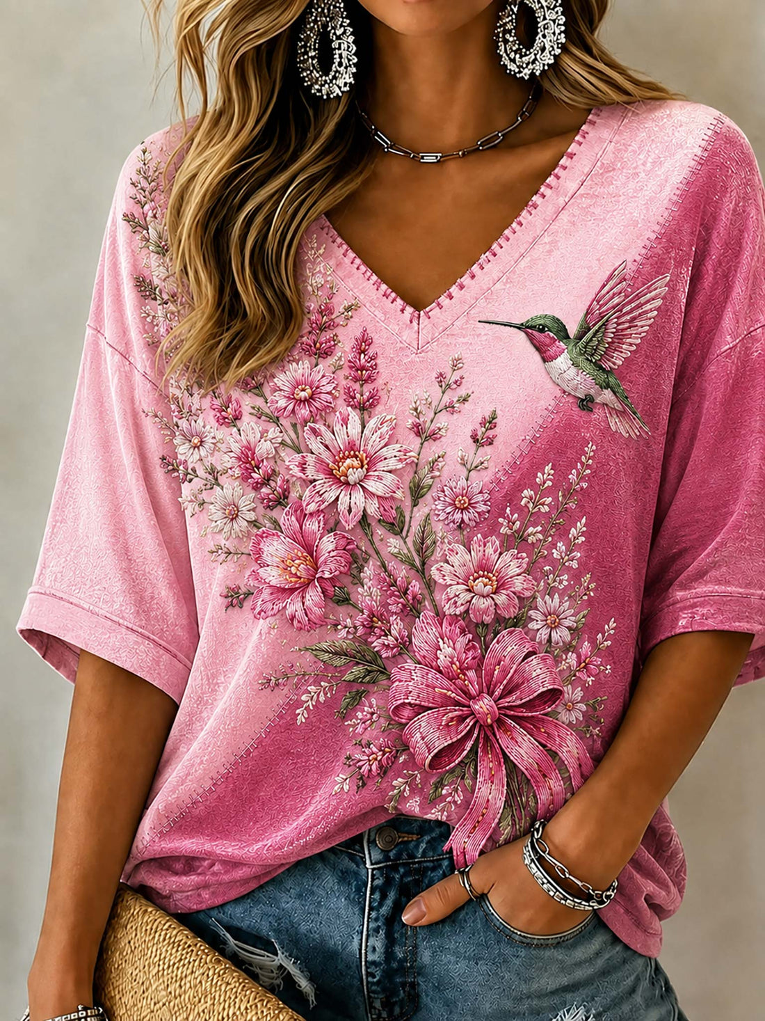 A woman wearing a pink short-sleeve V-neck top with a large floral and hummingbird graphic print, paired with blue jeans and silver jewelry.