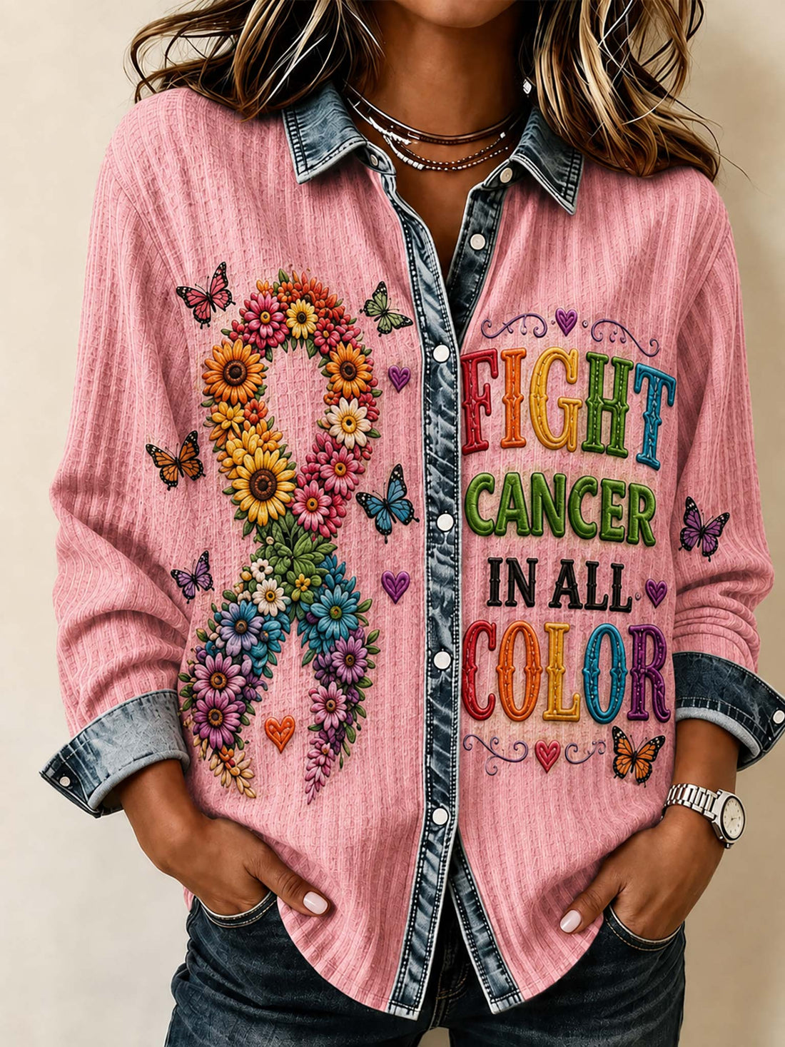 A woman wearing a pink long-sleeve button-up shirt with a denim collar and cuffs, featuring a colorful floral ribbon design on the left side and the text 'FIGHT CANCER IN ALL COLOR' on the right side.