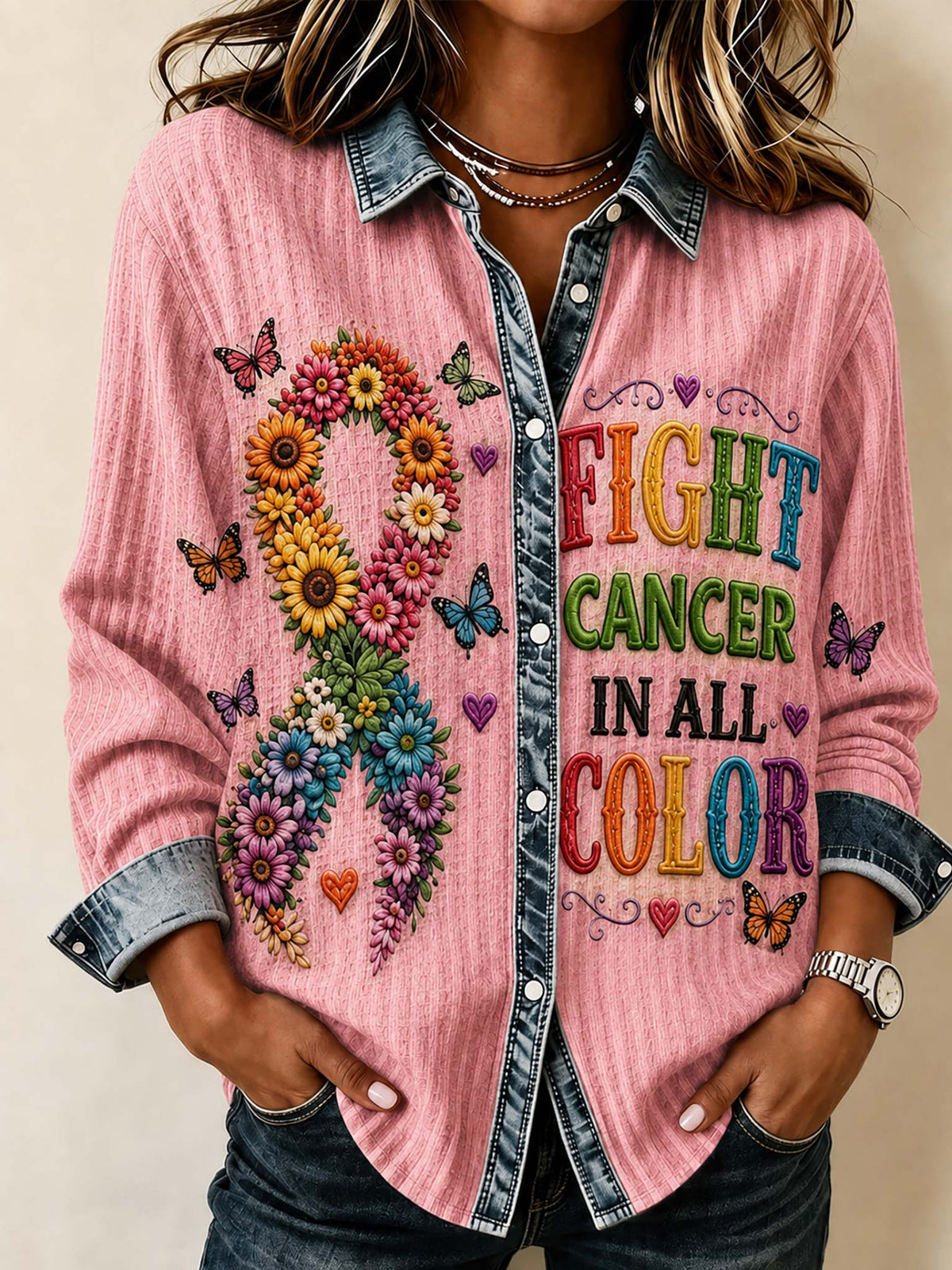 A woman wearing a pink long-sleeve button-up shirt with a denim collar and cuffs, featuring a colorful floral ribbon design on the left side and the text 'FIGHT CANCER IN ALL COLOR' on the right side.