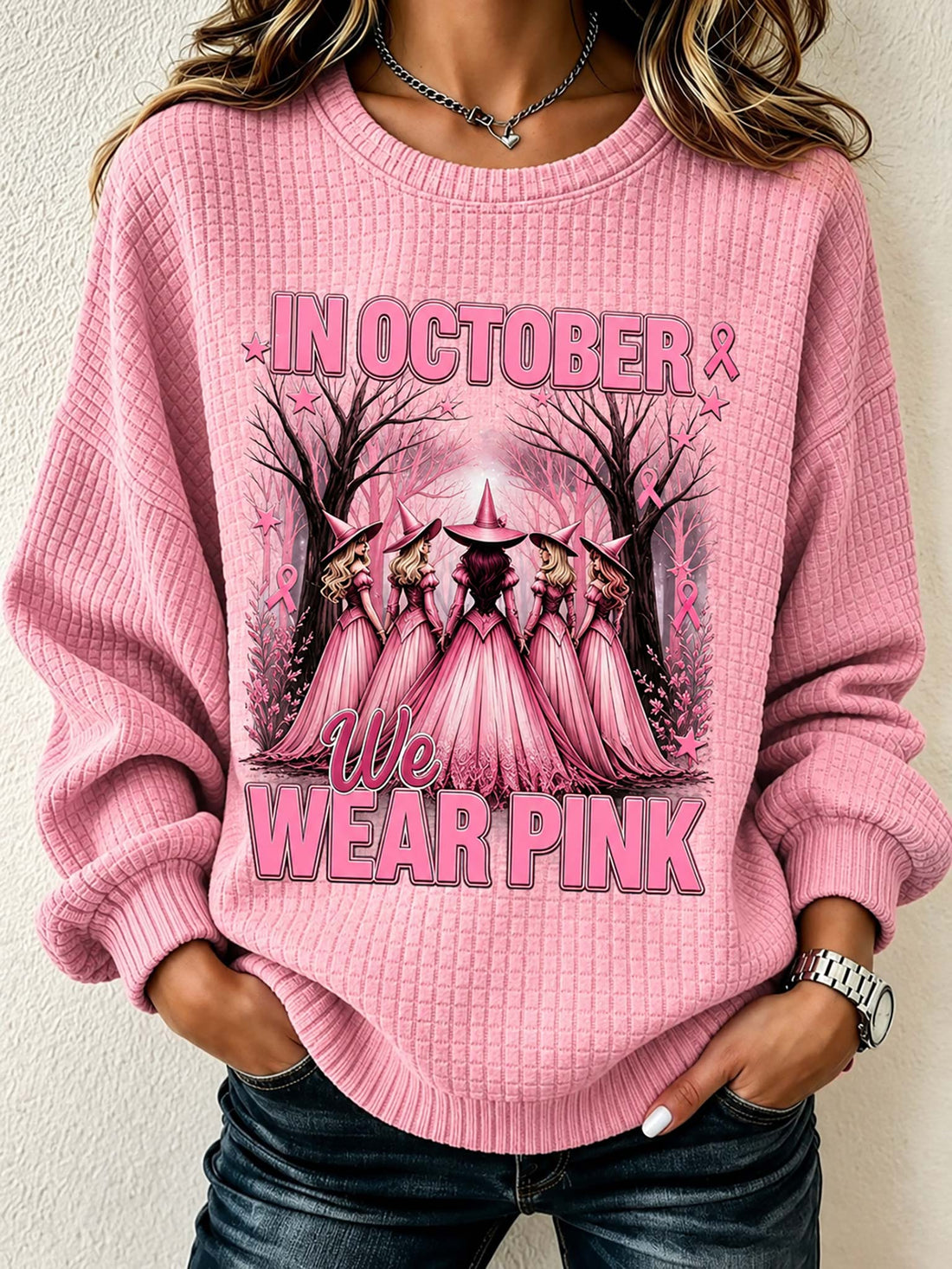 A woman wearing a pink waffle-knit crewneck sweatshirt with a 'We Wear Pink' graphic featuring pink-cloaked figures in a forest. The sweatshirt has long sleeves and ribbed cuffs and hem, paired with dark blue jeans.