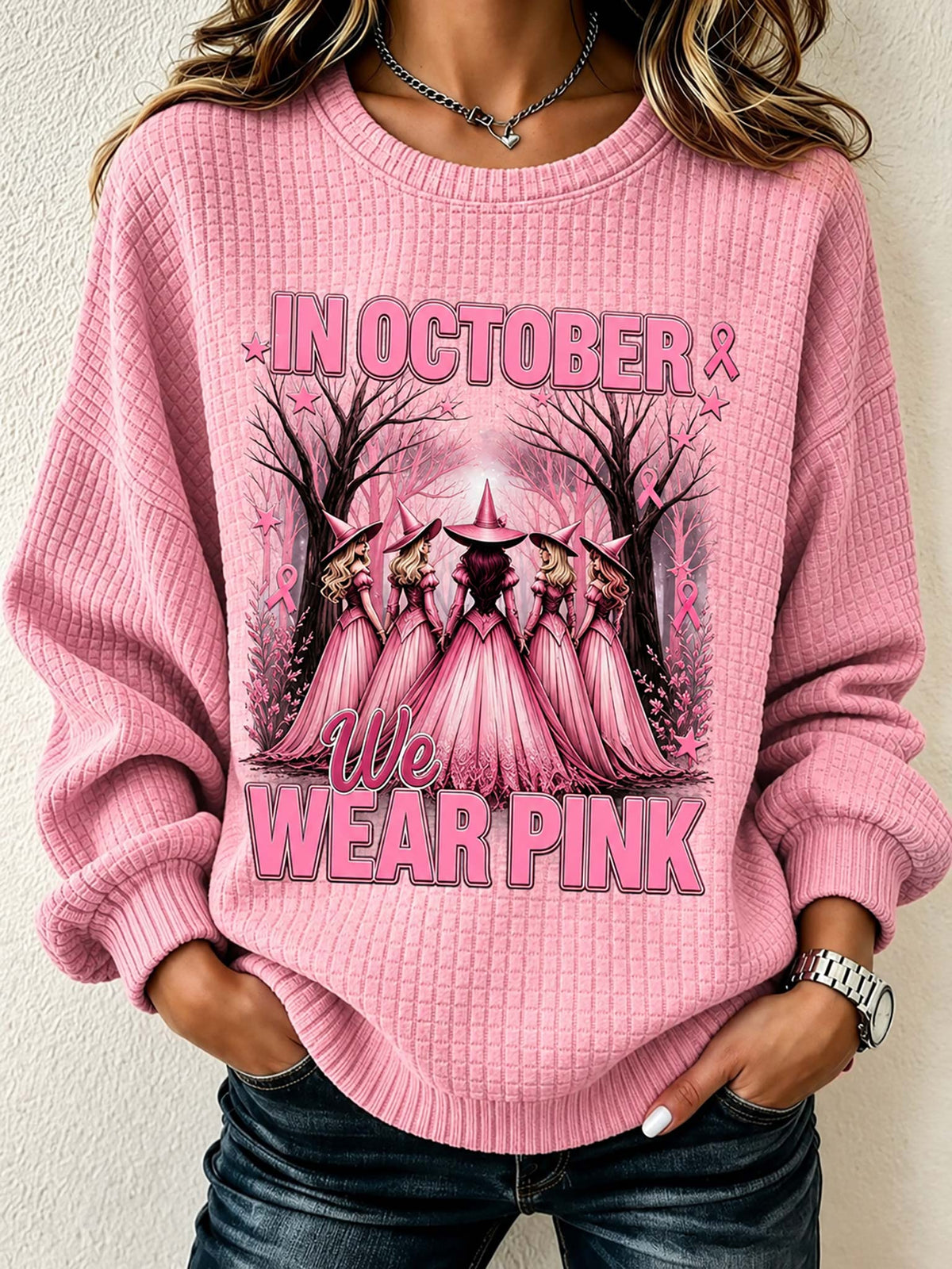 A woman wearing a pink waffle-knit crewneck sweatshirt with a 'We Wear Pink' graphic featuring pink-cloaked figures in a forest. The sweatshirt has long sleeves and ribbed cuffs and hem, paired with dark blue jeans.