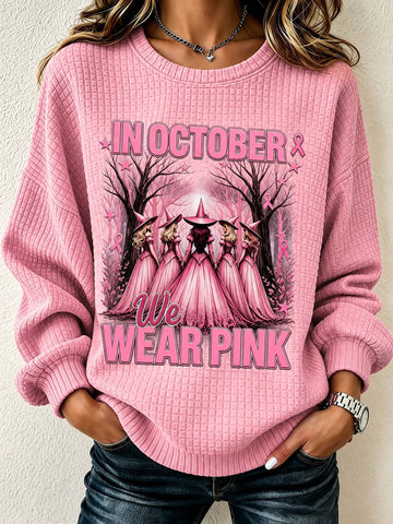 A woman wearing a pink waffle-knit crewneck sweatshirt with a 'We Wear Pink' graphic featuring pink-cloaked figures in a forest. The sweatshirt has long sleeves and ribbed cuffs and hem, paired with dark blue jeans.