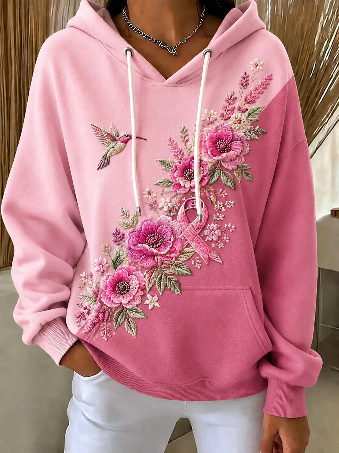 A pink pullover hoodie with white drawstrings, featuring a large floral and butterfly embroidery design on the front. The model is wearing white pants and a silver necklace, standing in an indoor setting.