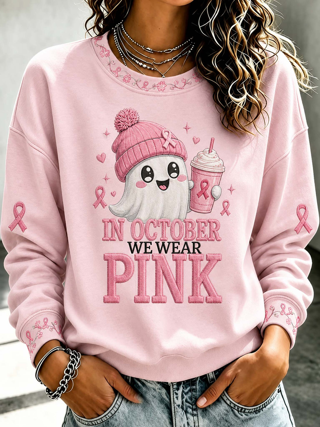 A woman wearing a light pink crewneck sweatshirt with a graphic that reads 'IN OCTOBER WE WEAR PINK' and features a cute ghost character in a pink beanie holding a pink drink, paired with blue jeans.