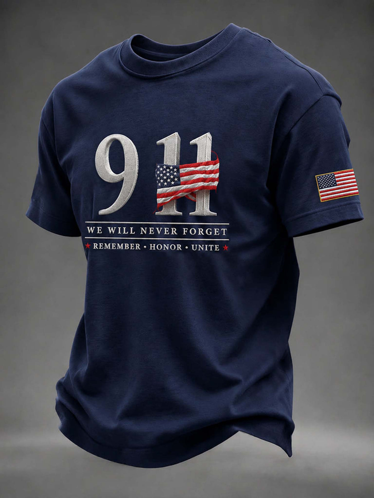 A navy blue short-sleeve t-shirt with a '911' memorial graphic on the chest, featuring an American flag pattern and the text 'WE WILL NEVER FORGET' and 'REMEMBER • HONOR • UNITE'. A small American flag is printed on the left sleeve, set against a dark gray studio background.
