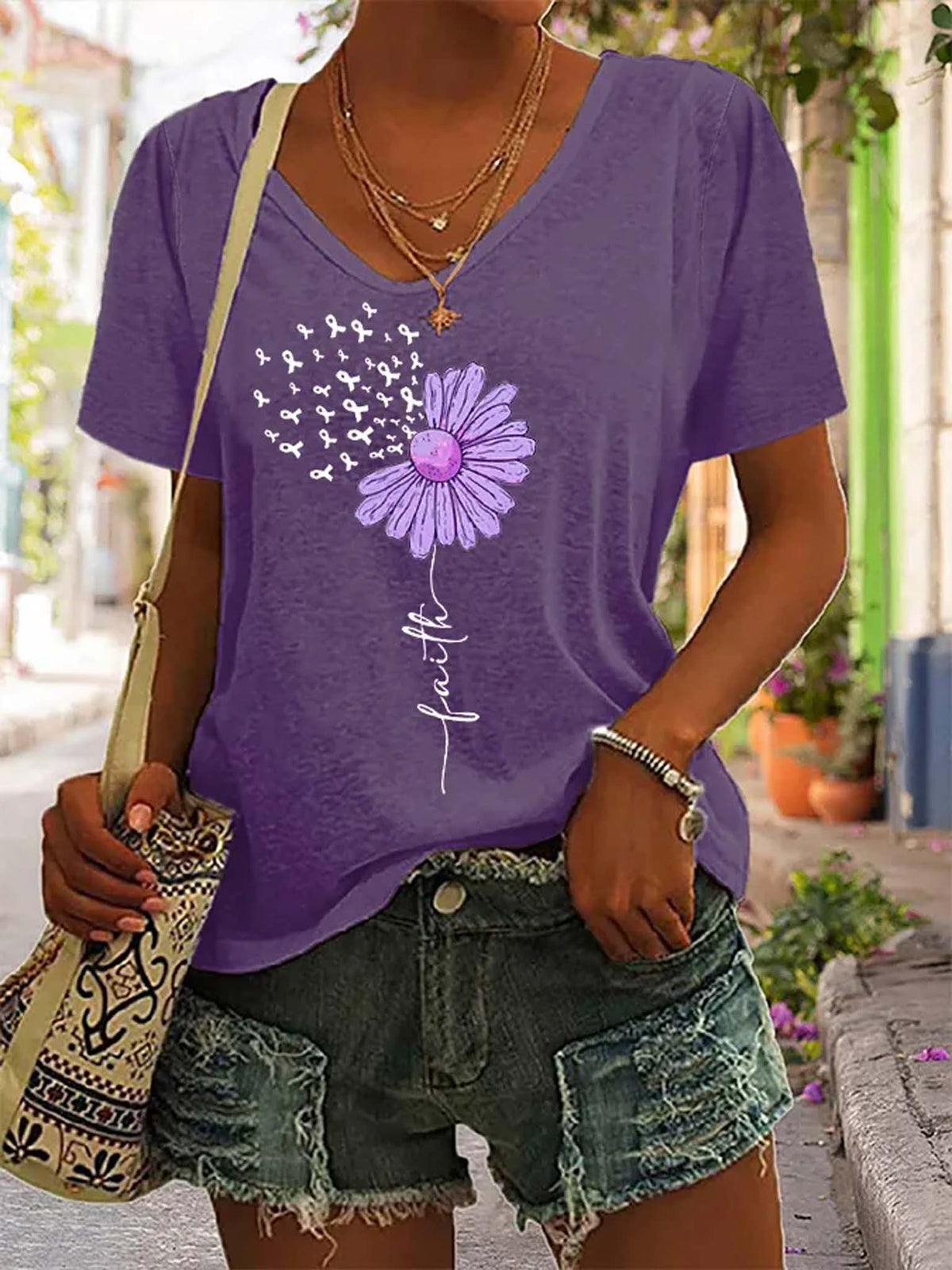 A woman wearing a purple short-sleeve V-neck t-shirt with a dandelion graphic and the word 'faith' printed on the front, paired with distressed denim shorts and carrying a patterned shoulder bag.
