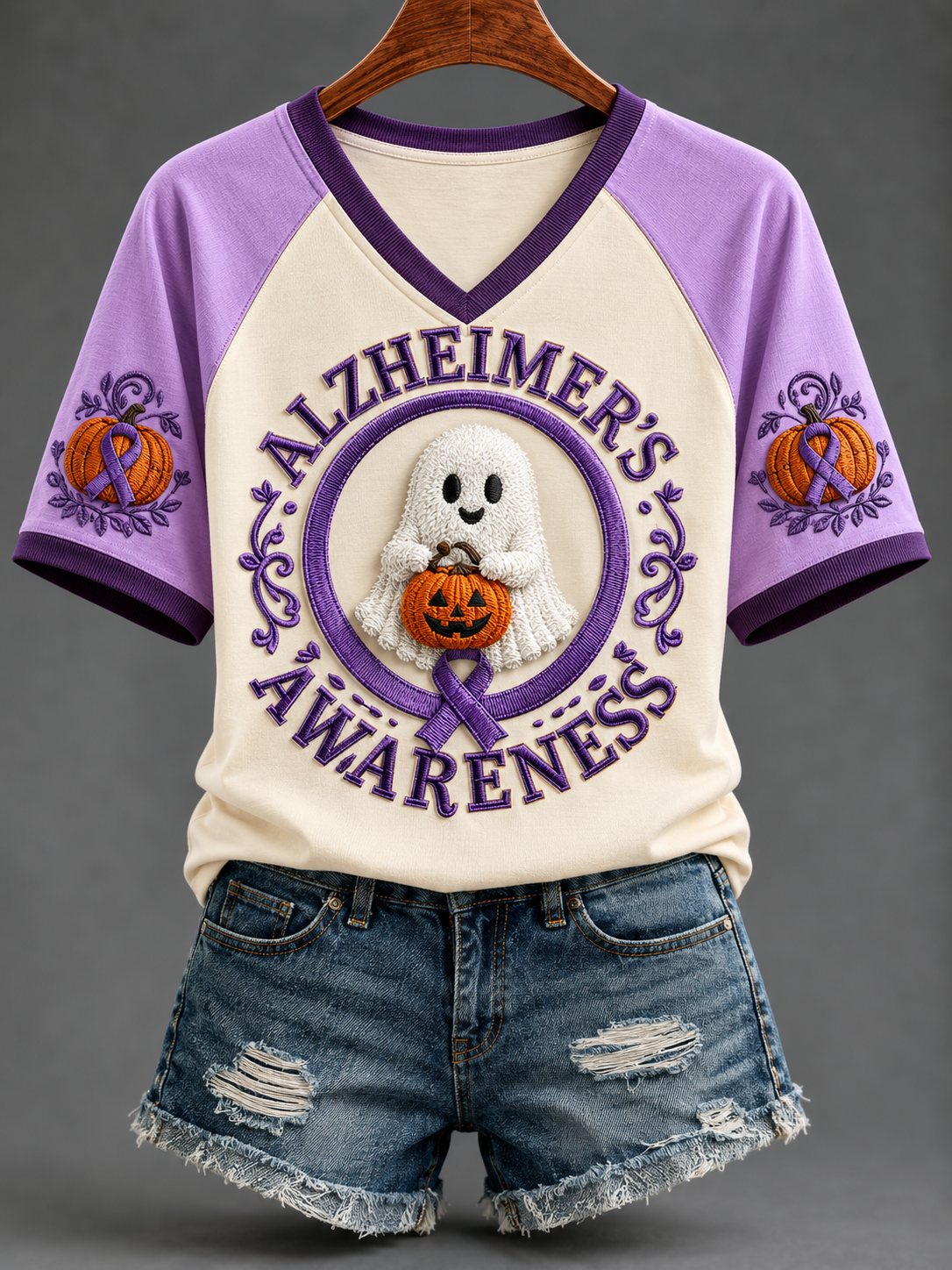 A raglan-style short-sleeve V-neck T-shirt with purple sleeves and collar trim, featuring a large front graphic that reads “ALZHEIMER’S AWARENESS” around a white ghost holding an orange pumpkin. The shirt is displayed on a hanger with distressed denim shorts against a neutral gray studio background.