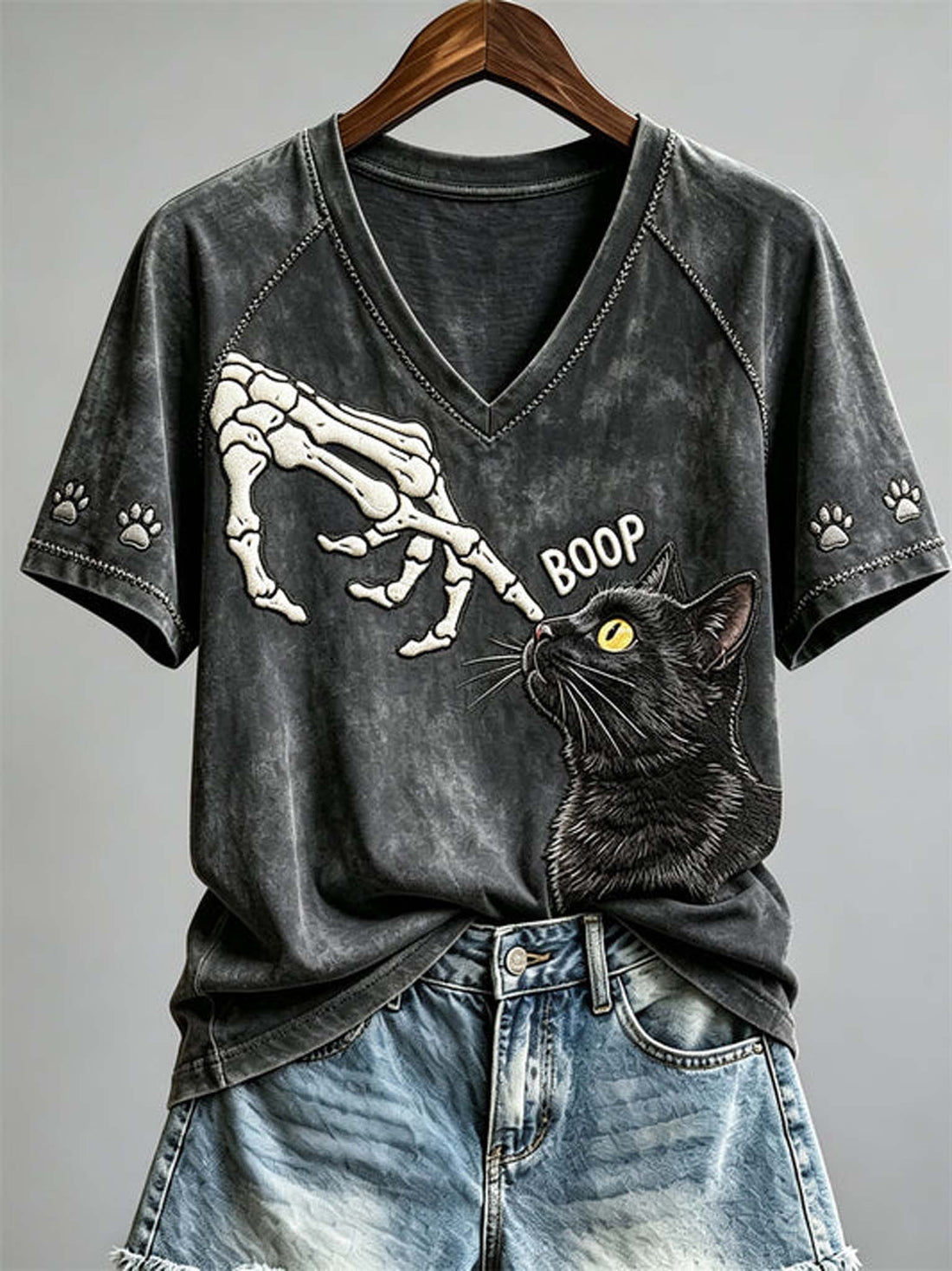 A dark gray washed V-neck t-shirt with a graphic of a skeleton hand biting a black cat's nose, with the word 'BOOP' and paw prints on the sleeves, shown hanging on a wooden hanger with blue denim shorts against a gray background.