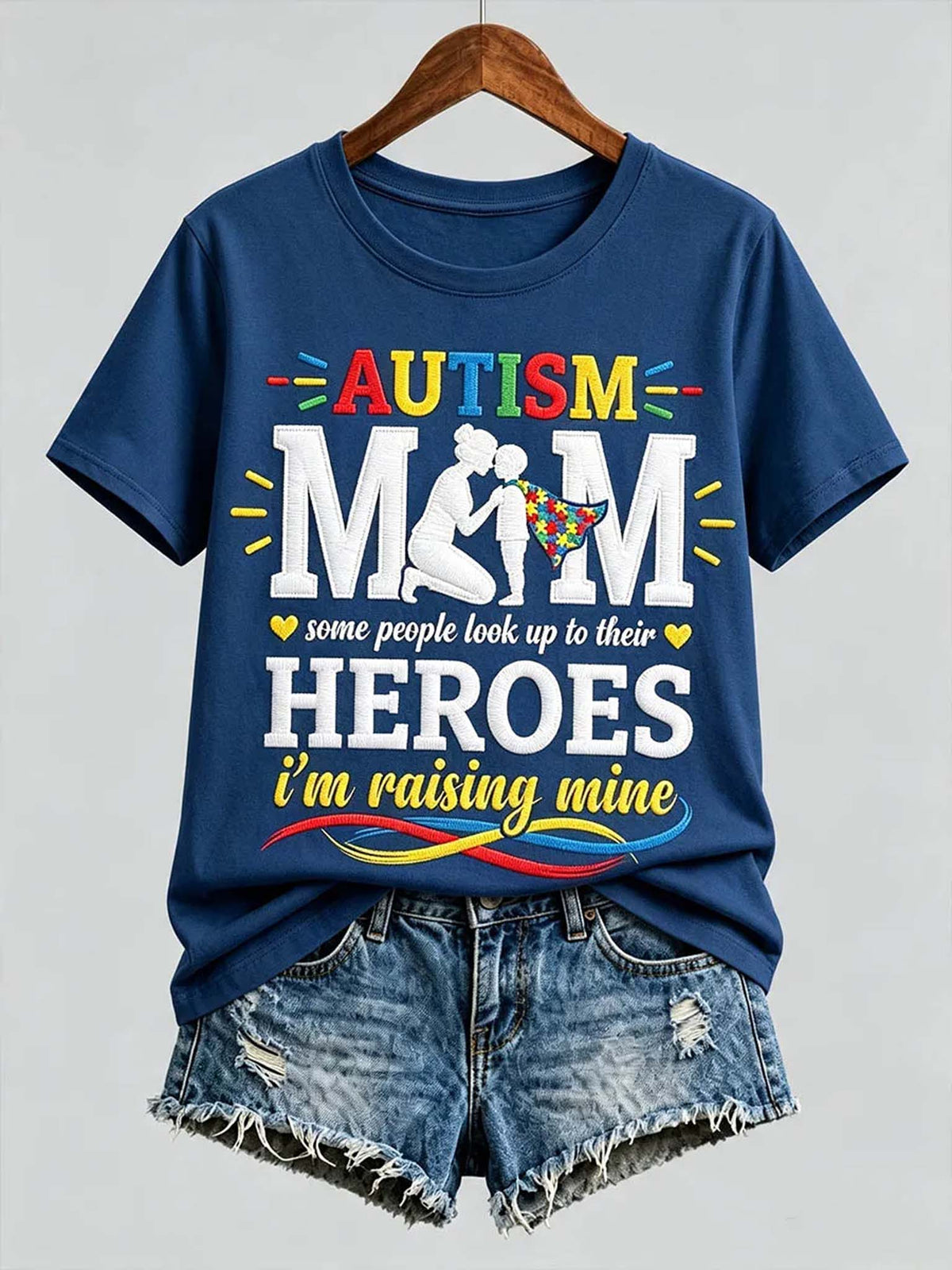A navy blue graphic t-shirt on a wooden hanger, styled with distressed denim shorts against a light gray background. The t-shirt features a colorful graphic that reads 'AUTISM MOM some people look up to their HEROES i'm raising mine' with a silhouette of a mother and child.