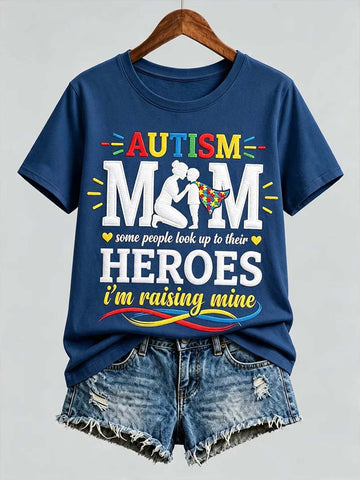 A navy blue graphic t-shirt on a wooden hanger, styled with distressed denim shorts against a light gray background. The t-shirt features a colorful graphic that reads 'AUTISM MOM some people look up to their HEROES i'm raising mine' with a silhouette of a mother and child.