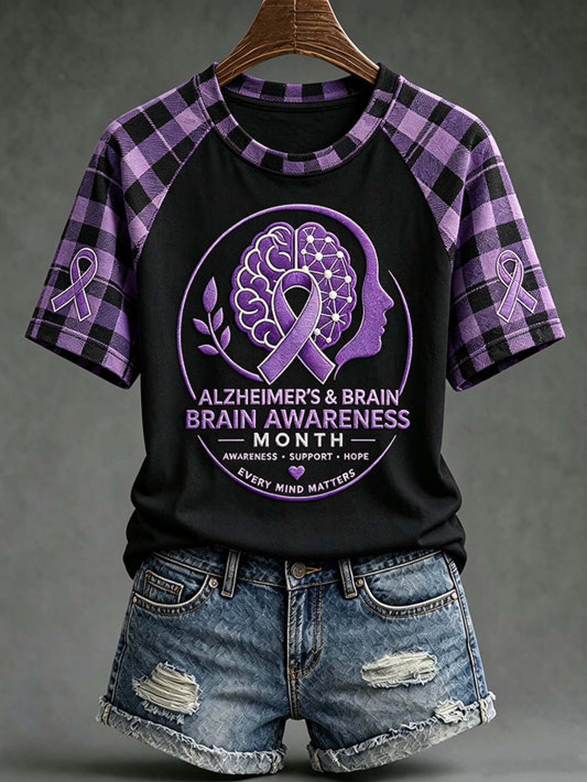 A women's short-sleeve raglan T-shirt with a black body and purple plaid sleeves, featuring a large Alzheimer's & Brain Awareness Month graphic on the front. The shirt is displayed on a wooden hanger against a dark gray background, styled with distressed denim shorts.