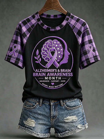 A women's short-sleeve raglan T-shirt with a black body and purple plaid sleeves, featuring a large Alzheimer's & Brain Awareness Month graphic on the front. The shirt is displayed on a wooden hanger against a dark gray background, styled with distressed denim shorts.