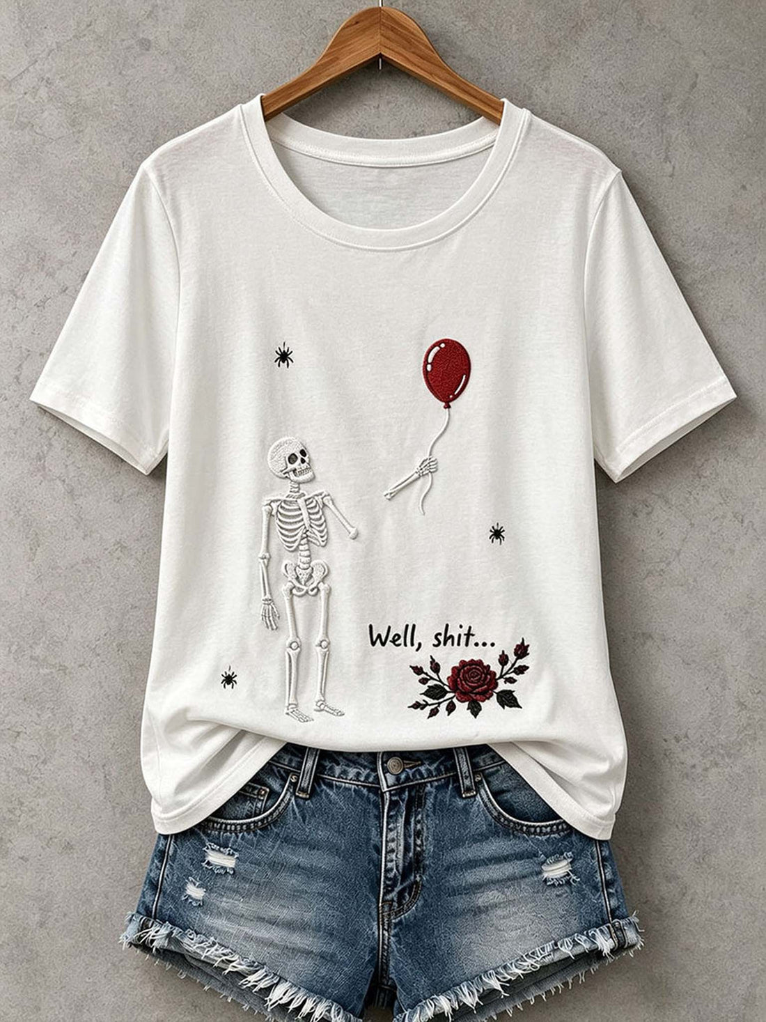 A white short-sleeve graphic T-shirt hanging on a wooden hanger against a gray textured wall, styled with distressed denim shorts. The shirt features a black line-art skeleton, a red balloon, the text 'Well, shit.