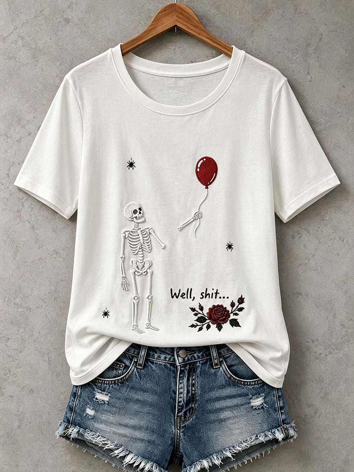 A white short-sleeve graphic T-shirt hanging on a wooden hanger against a gray textured wall, styled with distressed denim shorts. The shirt features a black line-art skeleton, a red balloon, the text 'Well, shit.