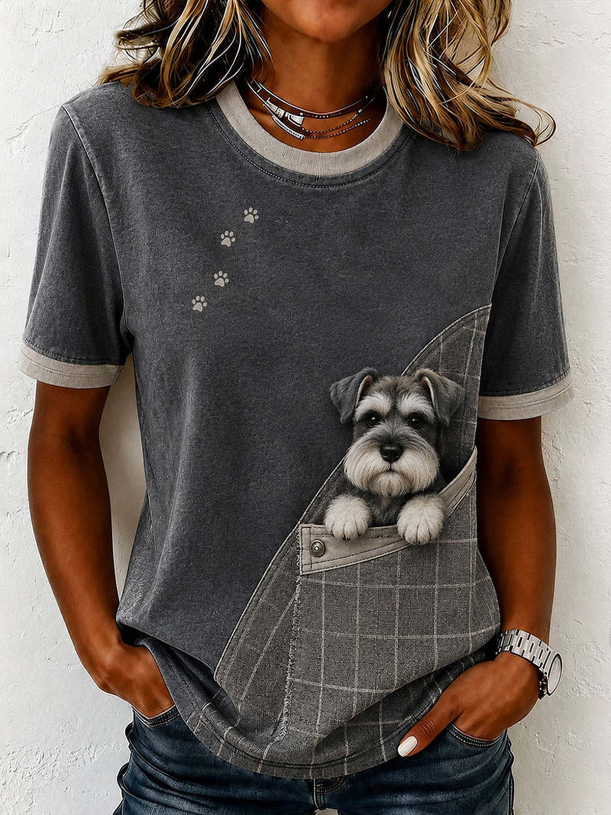 A woman wearing a dark gray short-sleeve t-shirt with a printed design of a gray and white schnauzer dog peeking out of a plaid pocket, set against a white textured wall.
