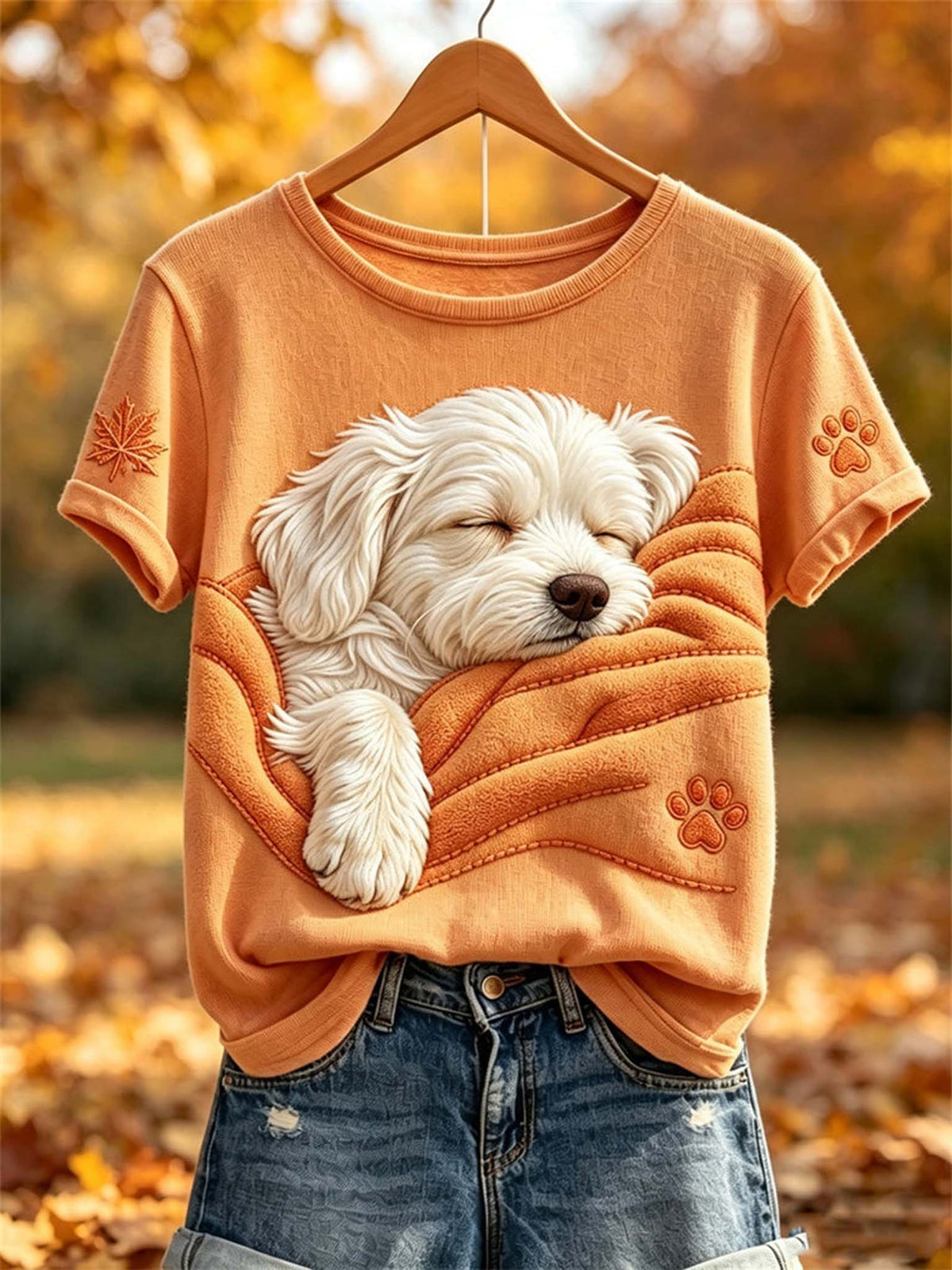 An orange short-sleeve t-shirt on a wooden hanger, featuring a large graphic of a sleeping white dog wrapped in an orange blanket. The shirt is styled with blue jeans, set against a blurred autumn background of fallen leaves and warm sunlight.