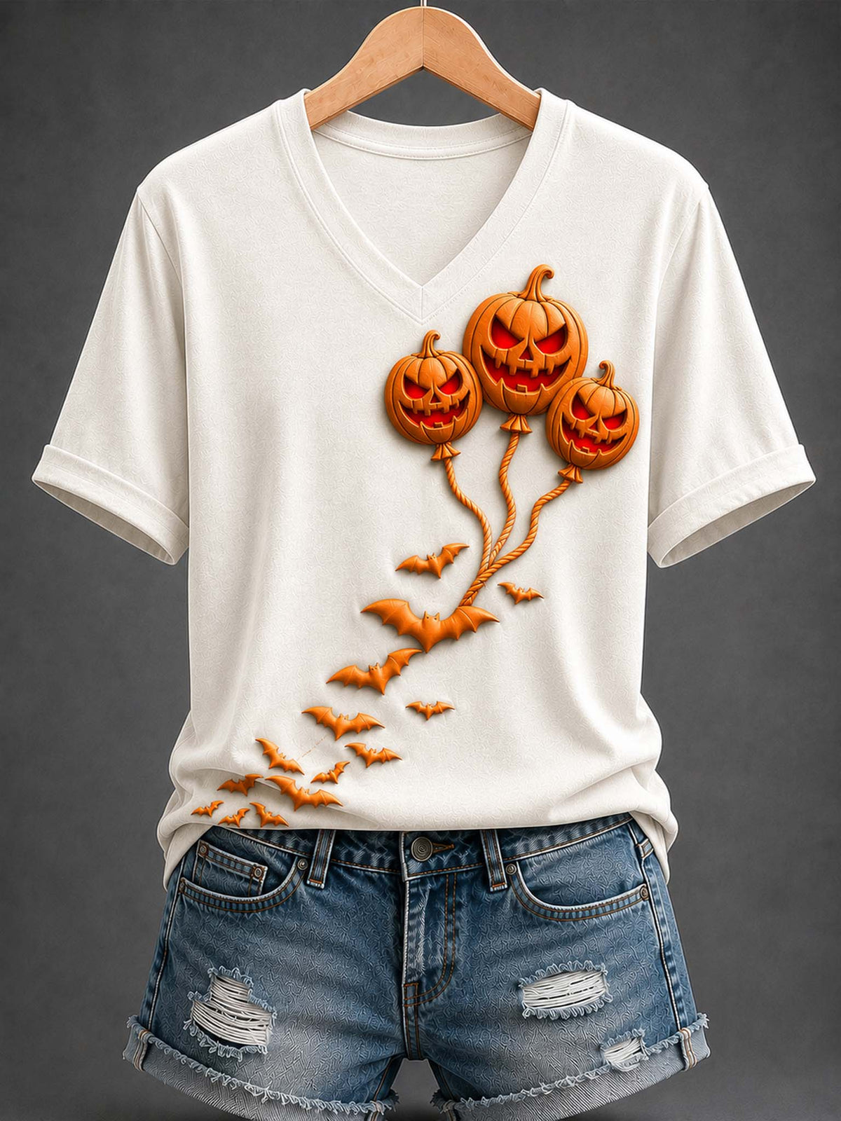A white V-neck t-shirt with a 3D-effect orange pumpkin balloon graphic and flying bats, styled on a wooden hanger with distressed denim shorts against a dark gray background.