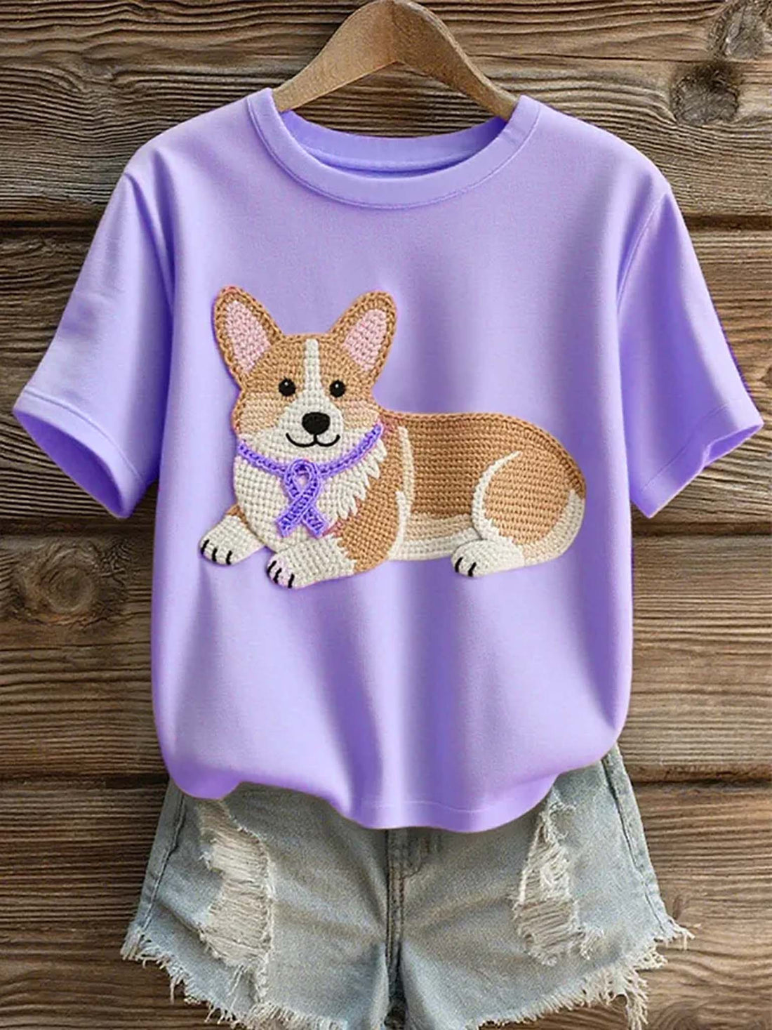 A lavender purple short-sleeve t-shirt with a crocheted corgi dog graphic on the front, styled with light-wash distressed denim shorts on a wooden hanger against a rustic wood plank background.