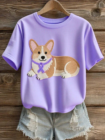 A lavender purple short-sleeve t-shirt with a crocheted corgi dog graphic on the front, styled with light-wash distressed denim shorts on a wooden hanger against a rustic wood plank background.