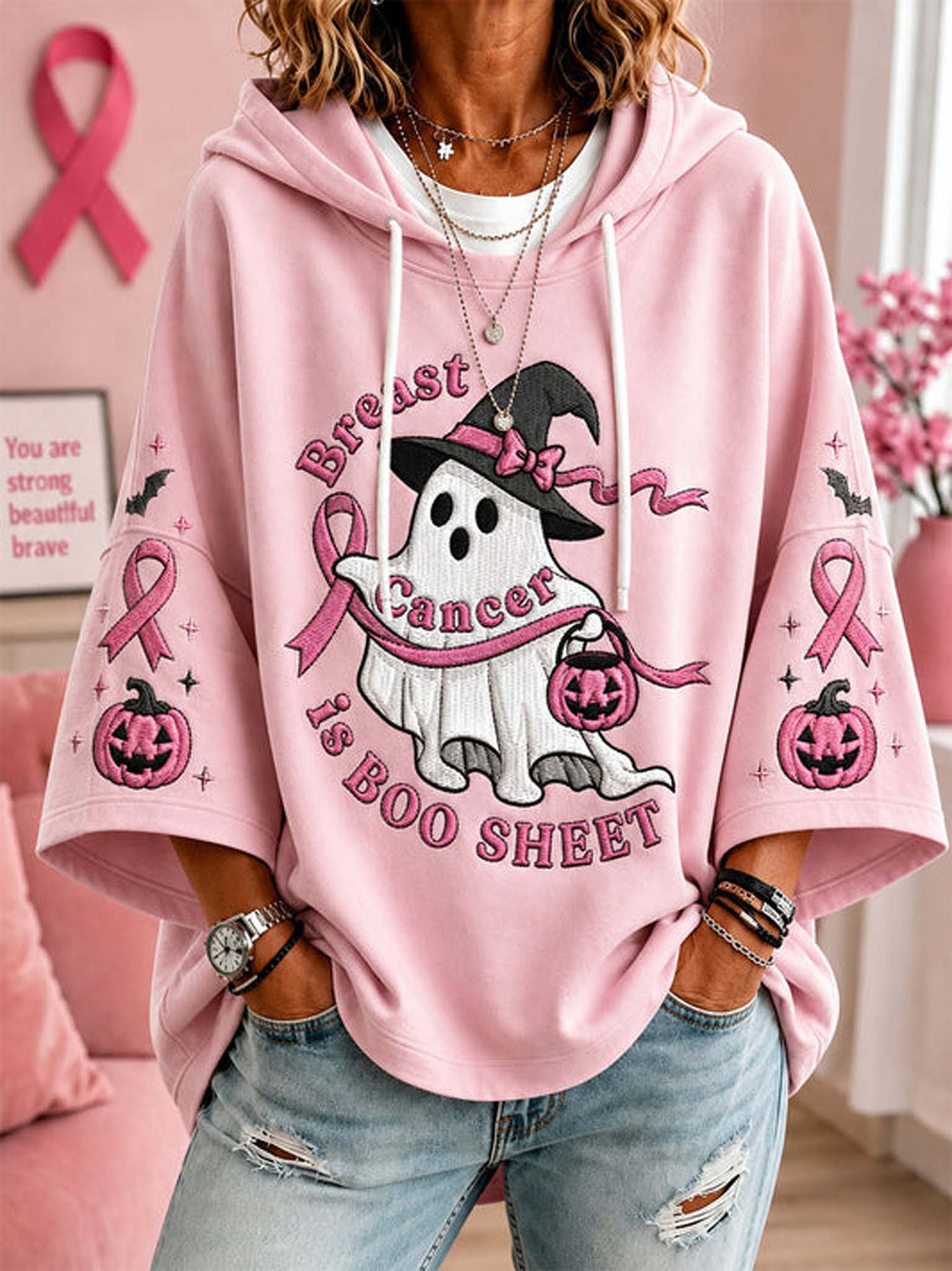 A woman wearing a pink oversized hoodie with a Halloween-themed breast cancer awareness graphic featuring a ghost in a witch hat, pumpkins, and pink ribbons, paired with distressed blue jeans.