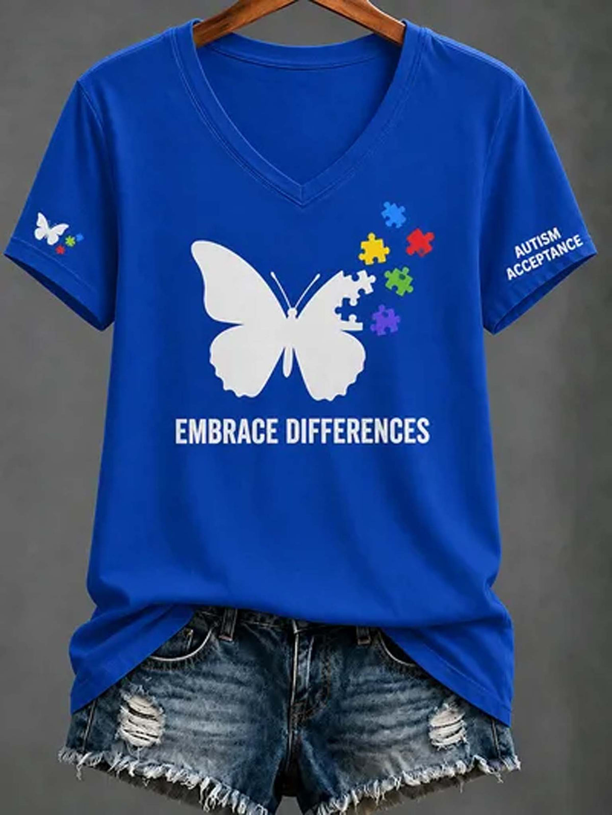 A royal blue V-neck t-shirt with a white butterfly graphic, colorful puzzle pieces, and the text 'EMBRACE DIFFERENCES' on the front, styled with distressed denim shorts on a hanger against a gray background.