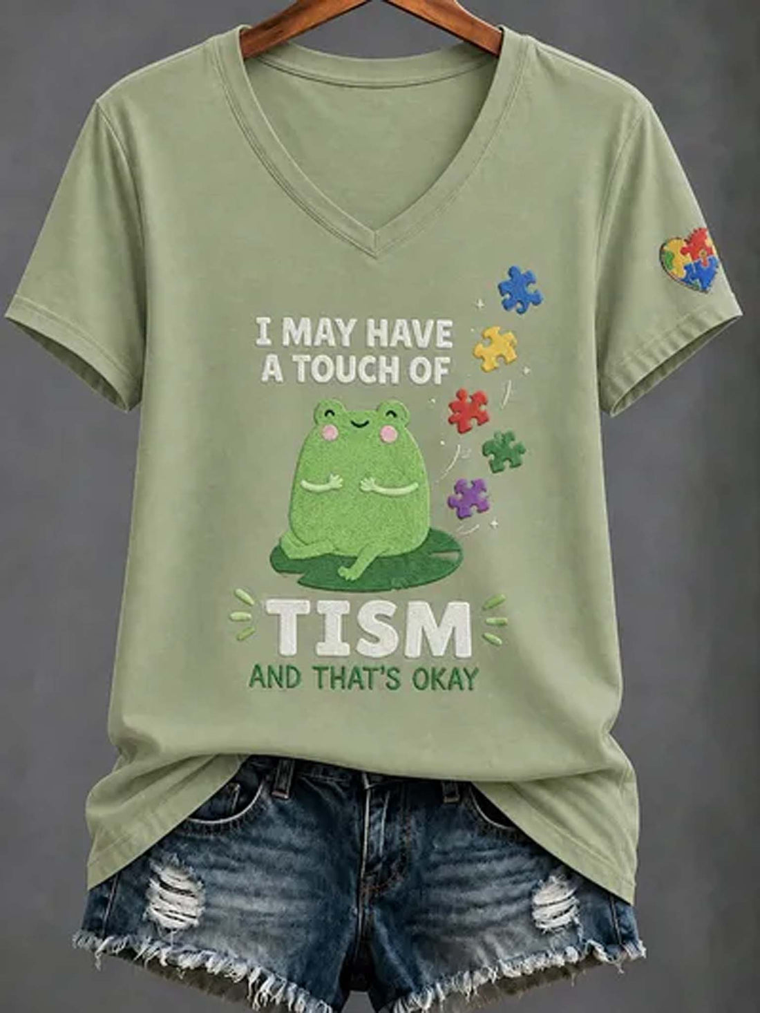 An olive green V-neck t-shirt with a graphic of a green frog and the text 'I MAY HAVE A TOUCH OF TISM AND THAT'S OKAY', styled on a hanger with distressed denim shorts against a dark gray background.