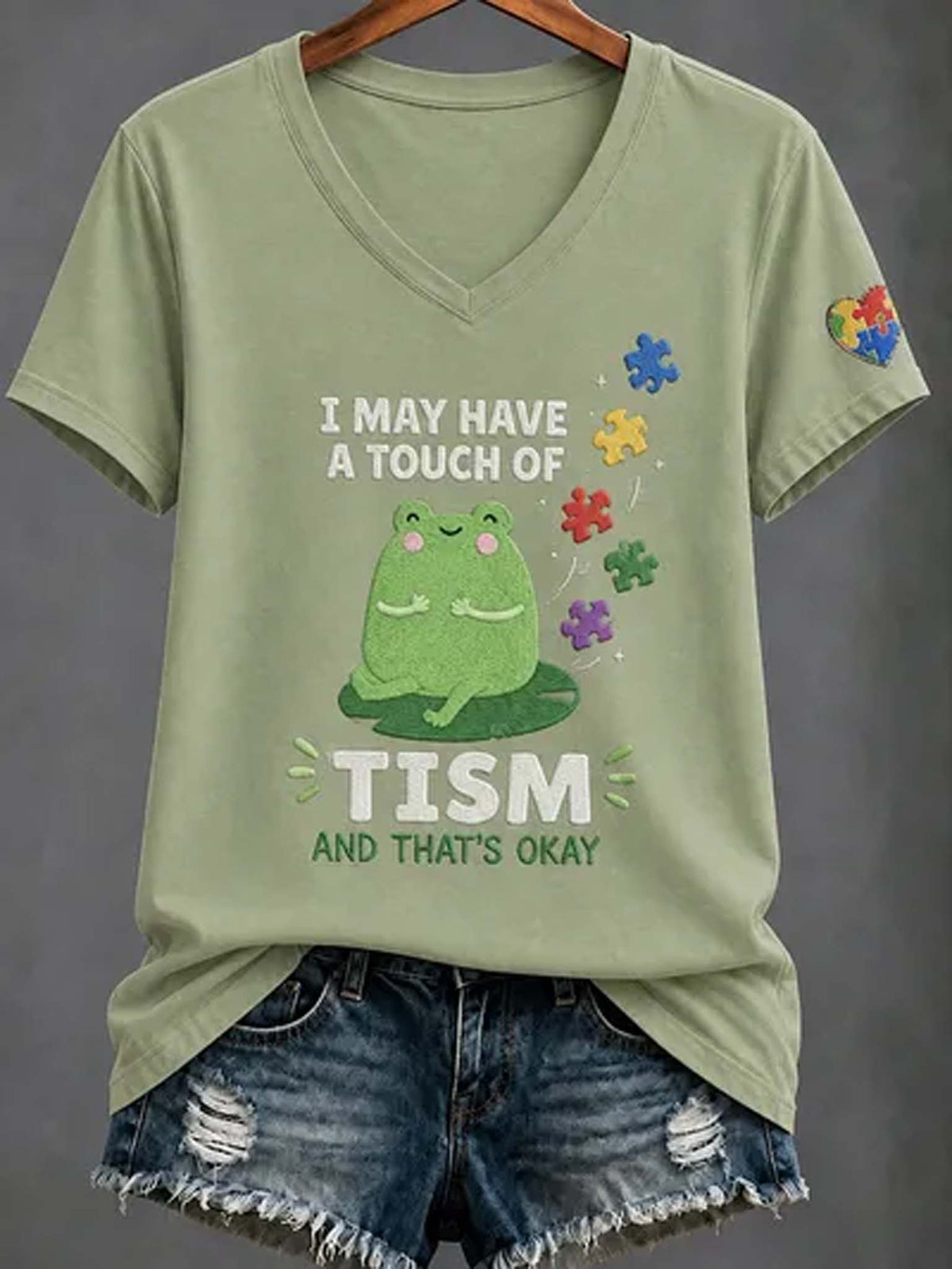 An olive green V-neck t-shirt with a graphic of a green frog and the text 'I MAY HAVE A TOUCH OF TISM AND THAT'S OKAY', styled on a hanger with distressed denim shorts against a dark gray background.