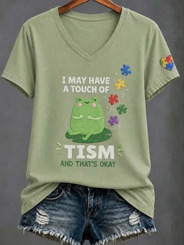 An olive green V-neck t-shirt with a graphic of a green frog and the text 'I MAY HAVE A TOUCH OF TISM AND THAT'S OKAY', styled on a hanger with distressed denim shorts against a dark gray background.