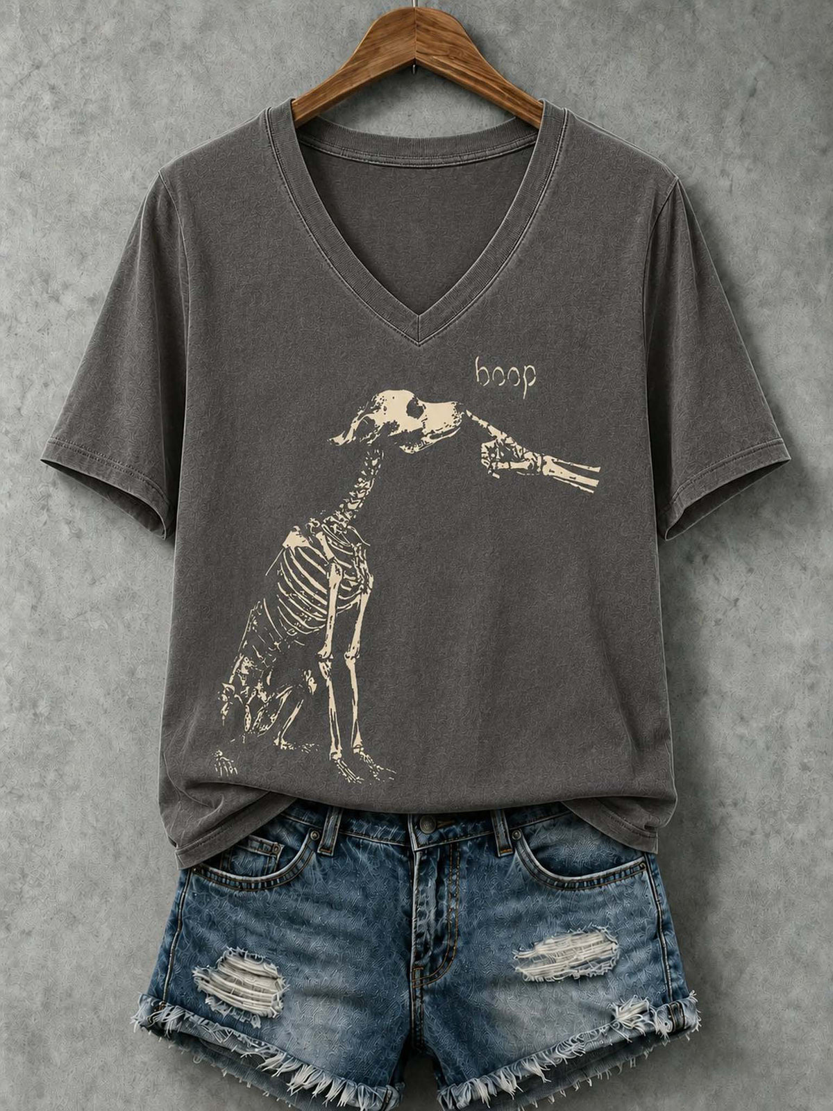 A dark gray v-neck t-shirt with a distressed beige graphic of a skeleton giraffe and the word 'hoop', styled on a wooden hanger with blue denim shorts against a textured gray background.