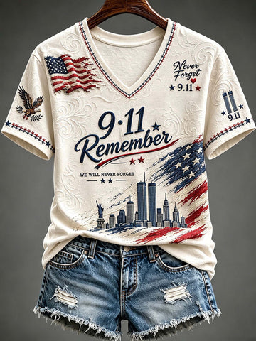 An off-white V-neck t-shirt with a patriotic 9/11 memorial graphic, styled with distressed denim shorts on a hanger against a dark gray background.