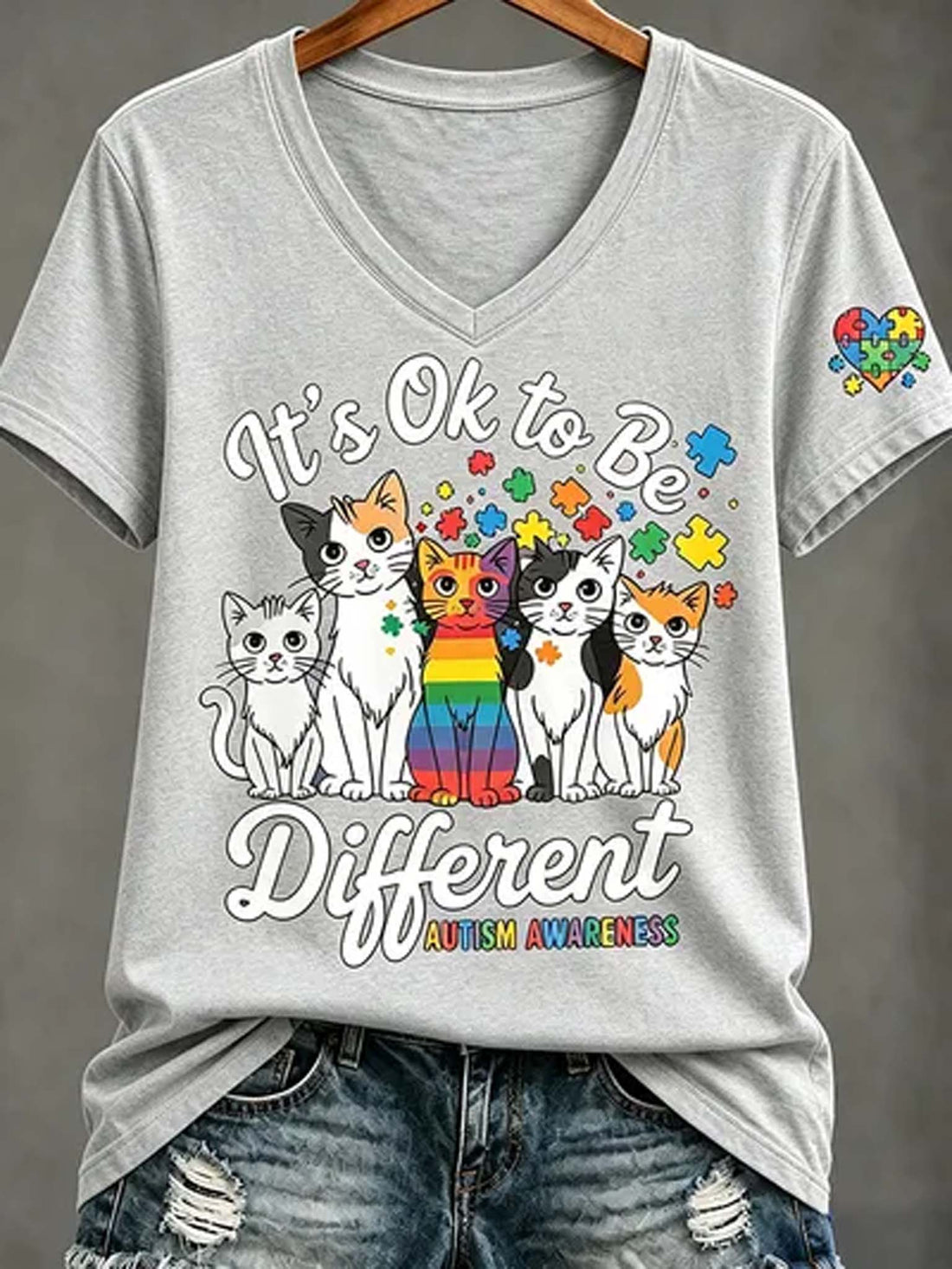 A light gray V-neck t-shirt with a colorful graphic of five cats and autism awareness puzzle pieces, styled on a wooden hanger with distressed denim jeans against a gray background.