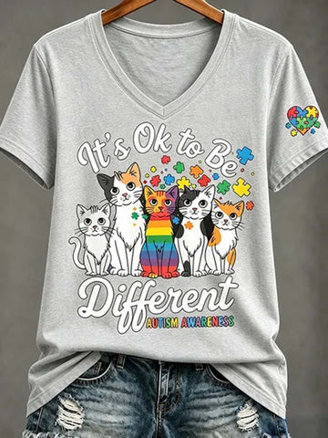 A light gray V-neck t-shirt with a colorful graphic of five cats and autism awareness puzzle pieces, styled on a wooden hanger with distressed denim jeans against a gray background.