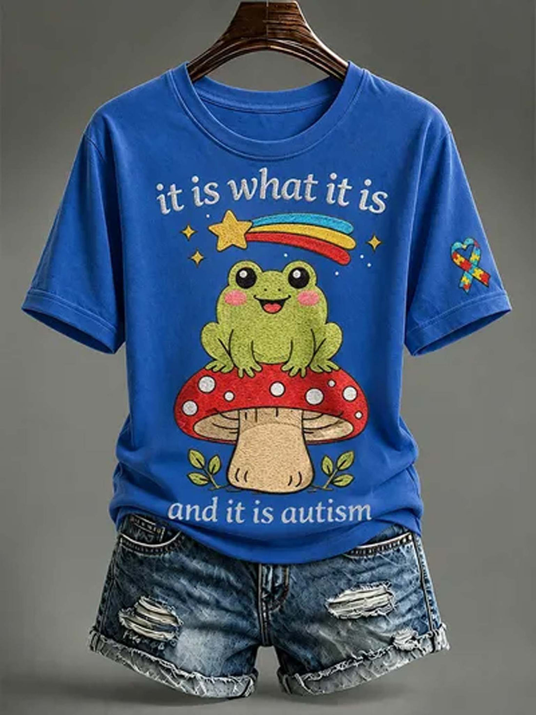 A blue graphic t-shirt displayed on a hanger with distressed denim shorts against a neutral gray background. The t-shirt features a cartoon green frog sitting on a red mushroom with the text 'it is what it is and it is autism' printed on the front.