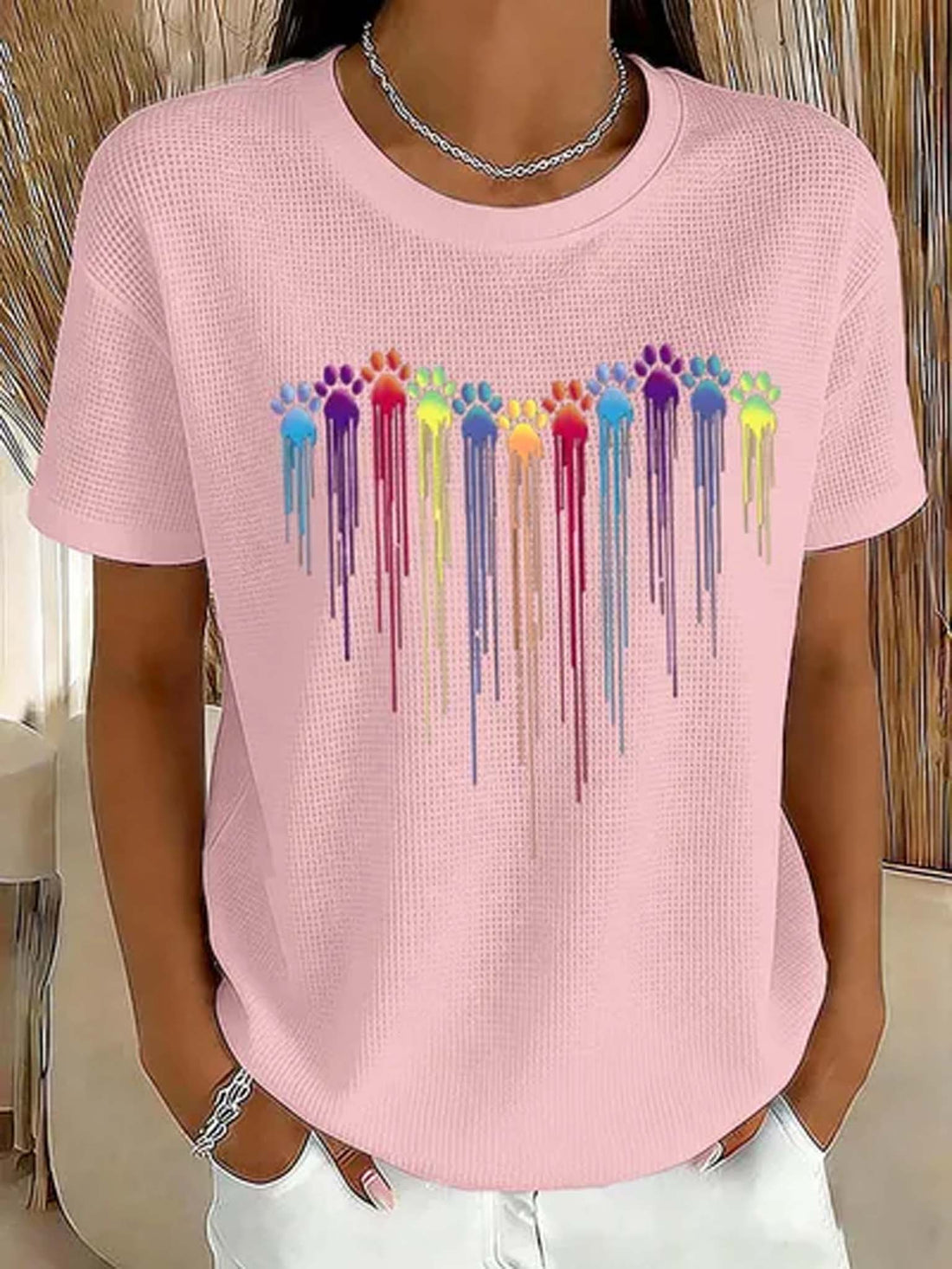 A woman wearing a light pink waffle-knit short-sleeve t-shirt with a colorful dripping paw print graphic across the chest, paired with white pants.