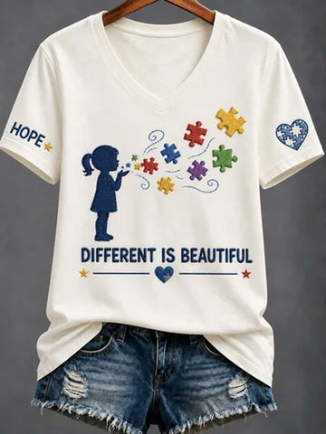 An off-white V-neck t-shirt with a Autism awareness graphic, styled with distressed denim shorts against a dark gray background. The graphic features a silhouette of a girl blowing puzzle pieces, with the text 'DIFFERENT IS BEAUTIFUL' and 'HOPE' on the sleeves.