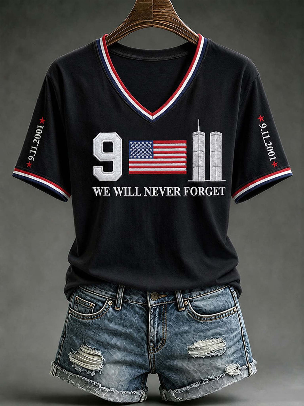 A black short-sleeve V-neck T-shirt with red, white, and blue trim, featuring a '9/11' memorial graphic with the American flag and the text 'WE WILL NEVER FORGET' on the front. The shirt is displayed on a wooden hanger with distressed denim shorts against a dark gray studio background.