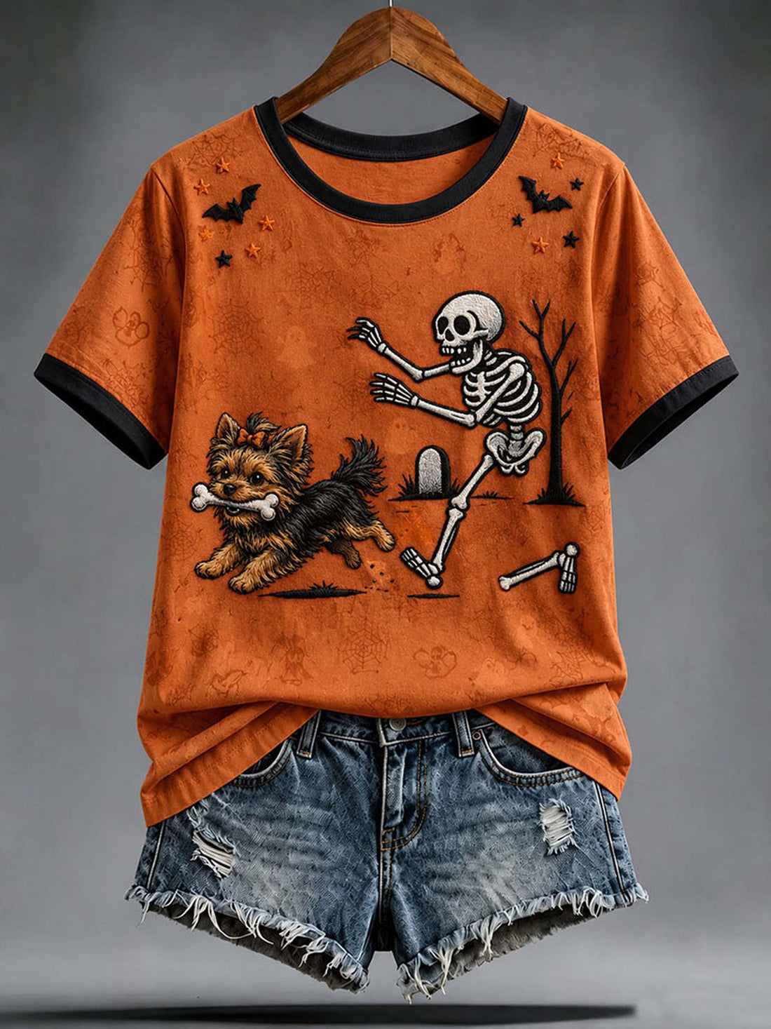 An orange short-sleeve ringer T-shirt with black trim and a Halloween graphic of a dancing skeleton and a small dog, styled with distressed denim shorts on a hanger against a gray studio background.