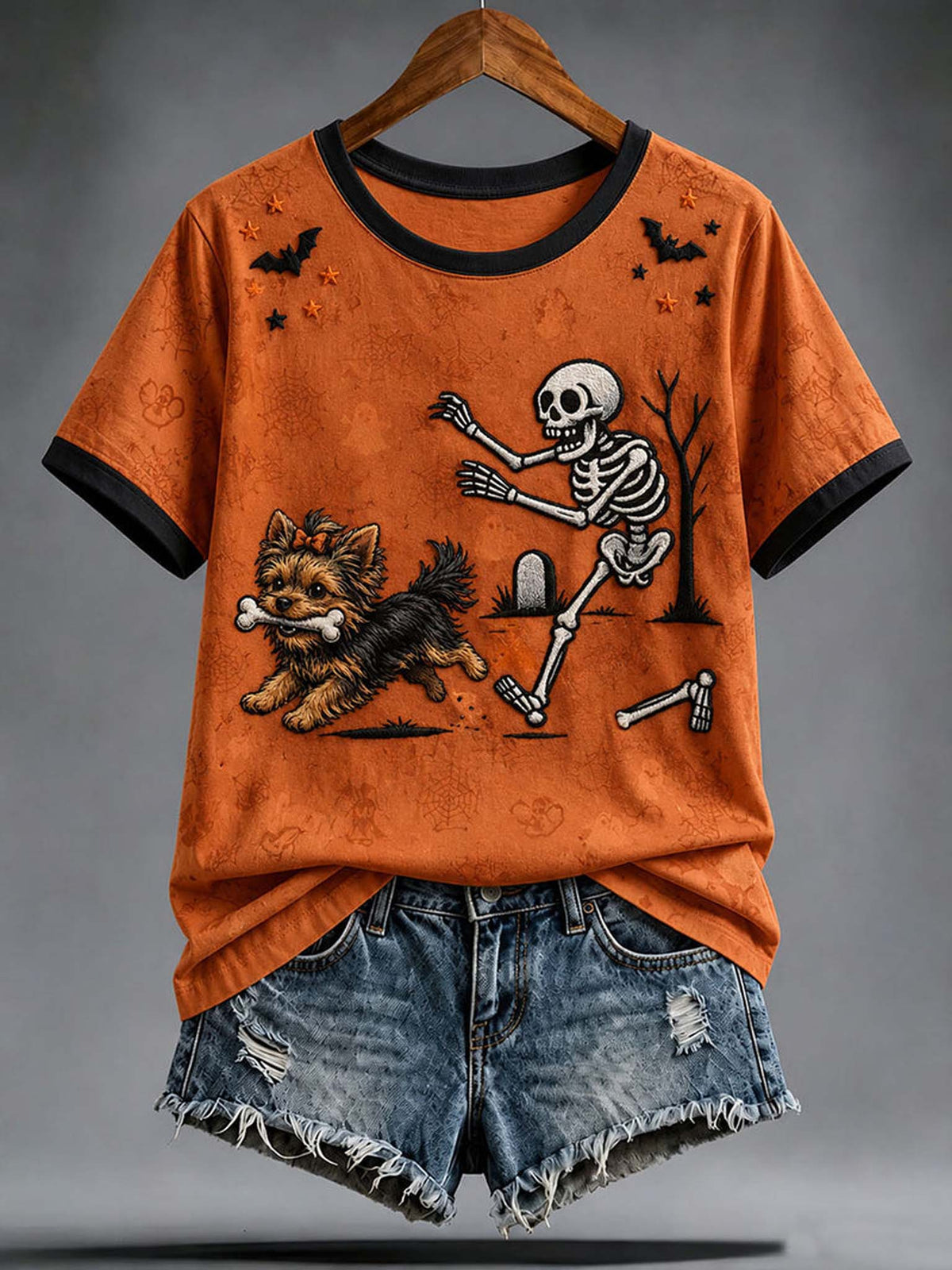 An orange short-sleeve ringer T-shirt with black trim and a Halloween graphic of a dancing skeleton and a small dog, styled with distressed denim shorts on a hanger against a gray studio background.