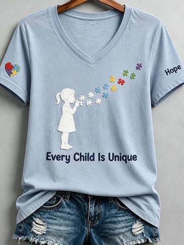 A light blue V-neck t-shirt with a graphic of a child blowing flower petals made of colorful puzzle pieces, with the text 'Every Child Is Unique' printed below. The t-shirt is styled with distressed denim shorts and hangs on a wooden hanger against a neutral gray background.