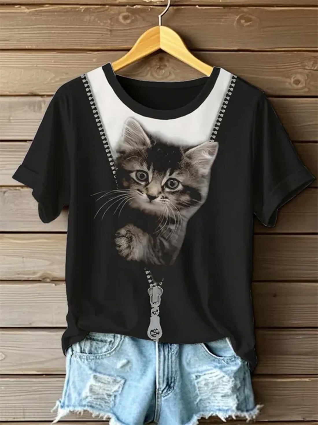 A black short-sleeve t-shirt with a graphic of a kitten hanging from a silver zipper on a white background, styled on a wooden hanger with light blue denim shorts against a wooden plank wall.