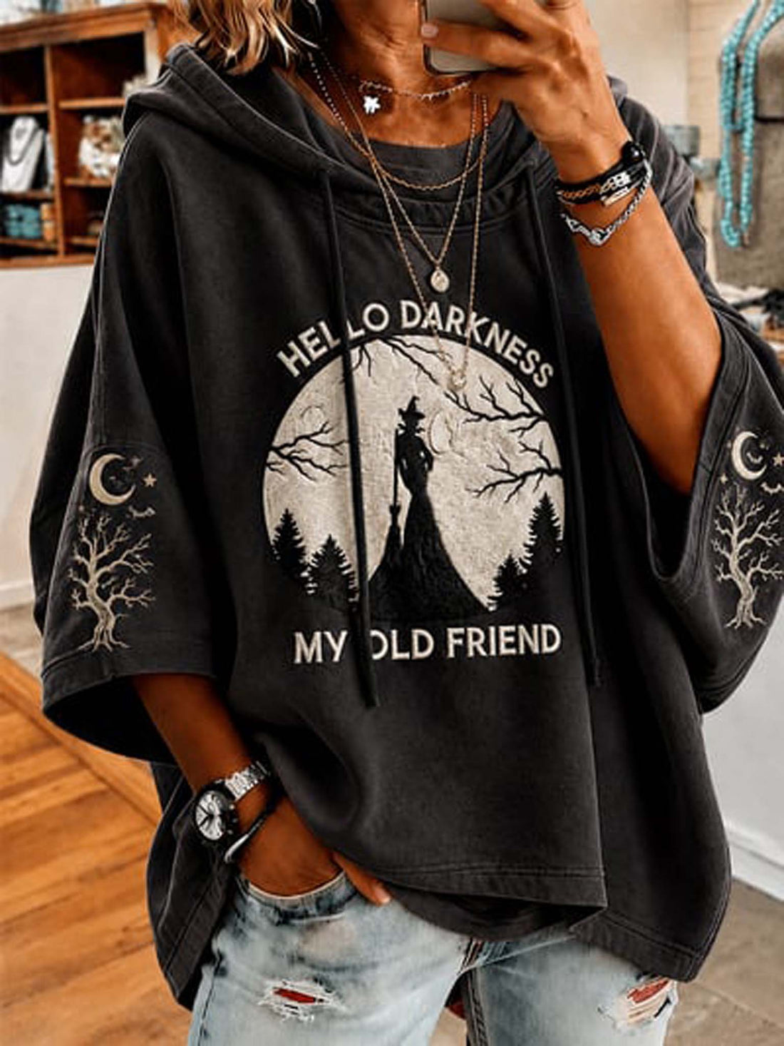 A person wearing a dark charcoal gray oversized hoodie with a distressed graphic that reads 'HELLO DARKNESS MY OLD FRIEND' around a moonlit forest scene. The hoodie has sleeve prints of a crescent moon and tree.