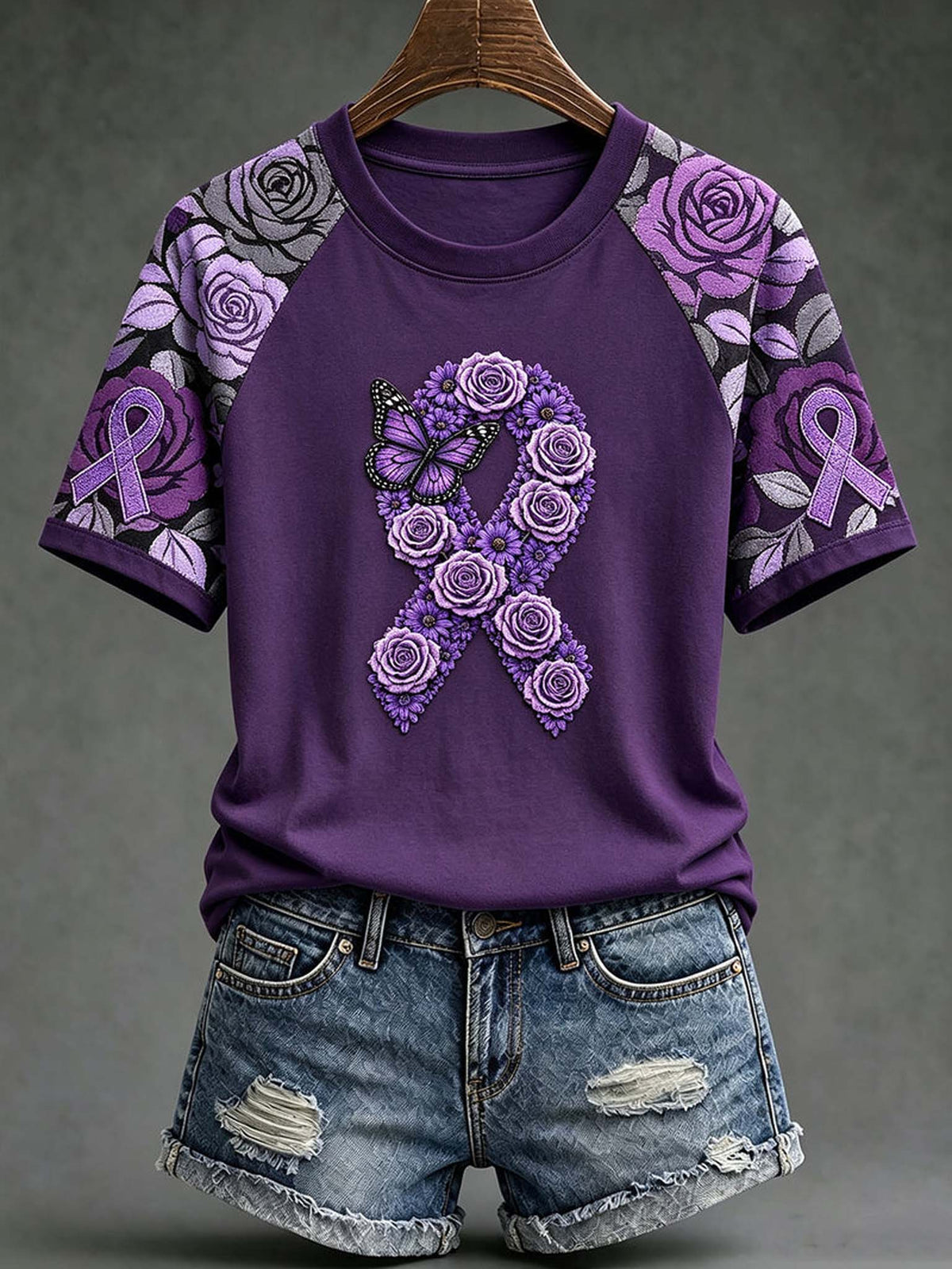 A purple short-sleeve raglan T-shirt with a large purple rose and ribbon graphic on the front, styled with distressed denim shorts on a hanger against a dark gray background.