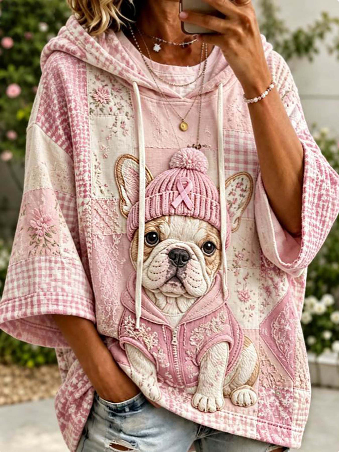 A person wearing a pink and cream patchwork hoodie with a large embroidered French Bulldog design on the front, paired with light blue distressed jeans, photographed outdoors.