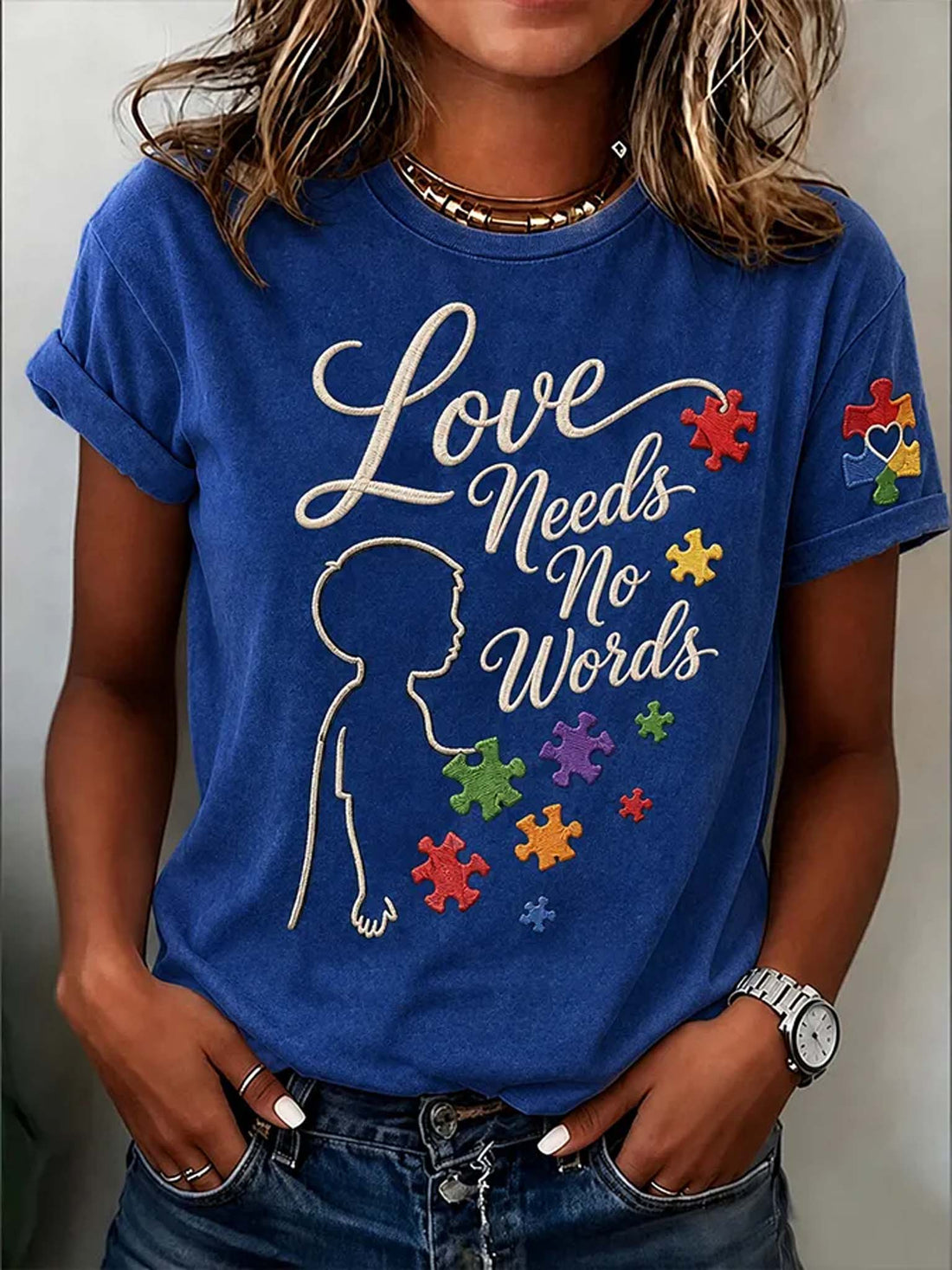 A woman wearing a royal blue graphic t-shirt with the text 'Love Needs No Words' in white script, accompanied by a white silhouette of a child and colorful autism awareness puzzle pieces. She is also wearing blue jeans and a silver watch.