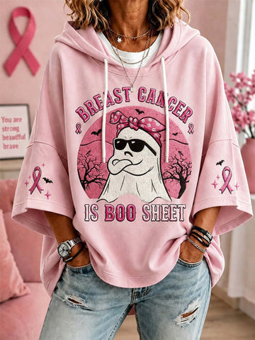 A woman wearing a pink graphic hoodie with a ghost wearing sunglasses and a pink bow, surrounded by Halloween-themed elements, with the text 'BREAST CANCER IS BOO SHEET' printed on the front. She is also wearing distressed blue jeans.