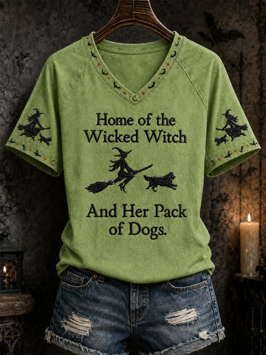 A sage green raglan V-neck t-shirt with a black graphic print reading 'Home of the Wicked Witch And Her Pack of Dogs' with witch and dog illustrations, styled on a hanger with distressed denim shorts against a dark, atmospheric Halloween background.