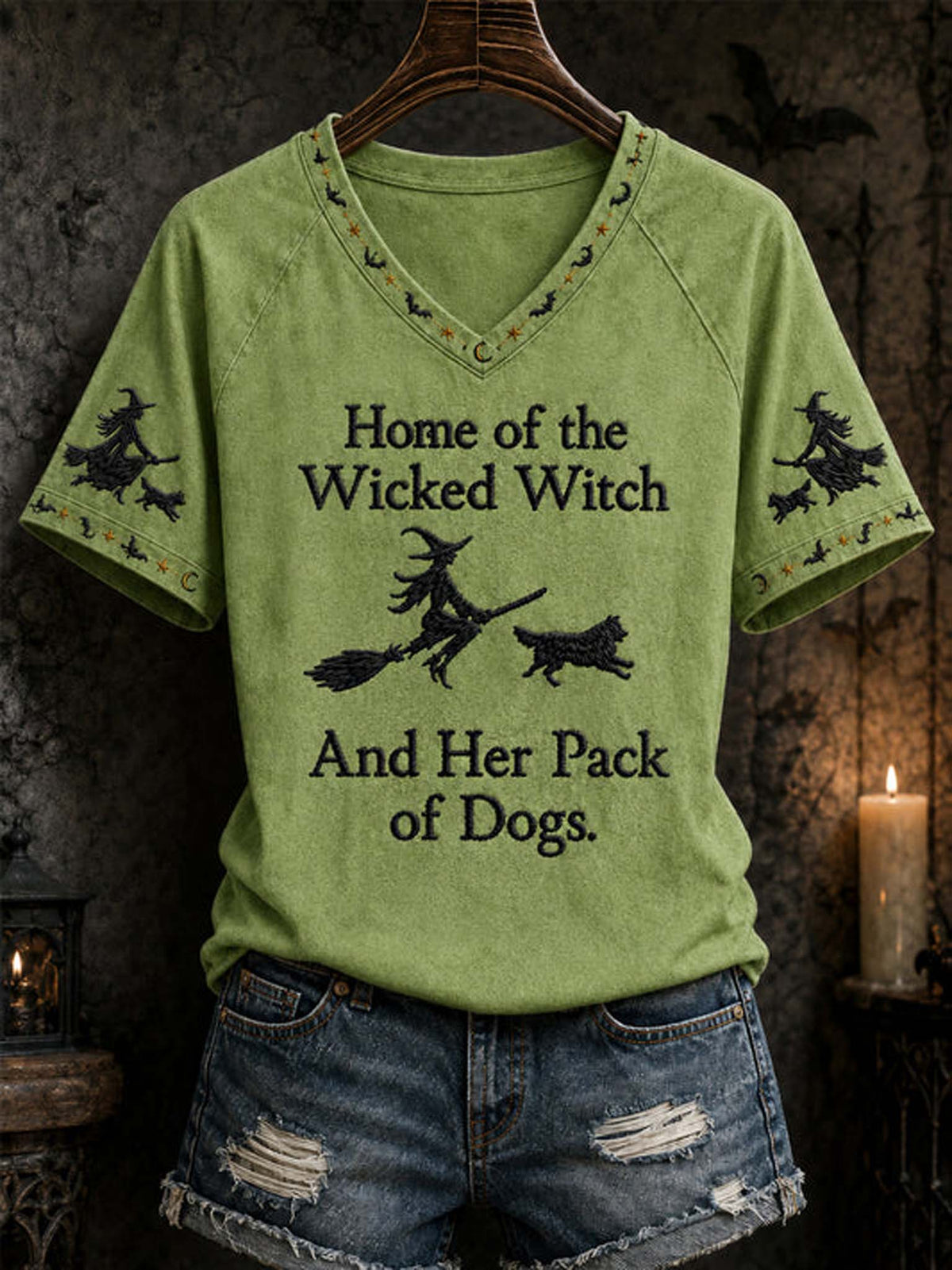 A sage green raglan V-neck t-shirt with a black graphic print reading 'Home of the Wicked Witch And Her Pack of Dogs' with witch and dog illustrations, styled on a hanger with distressed denim shorts against a dark, atmospheric Halloween background.